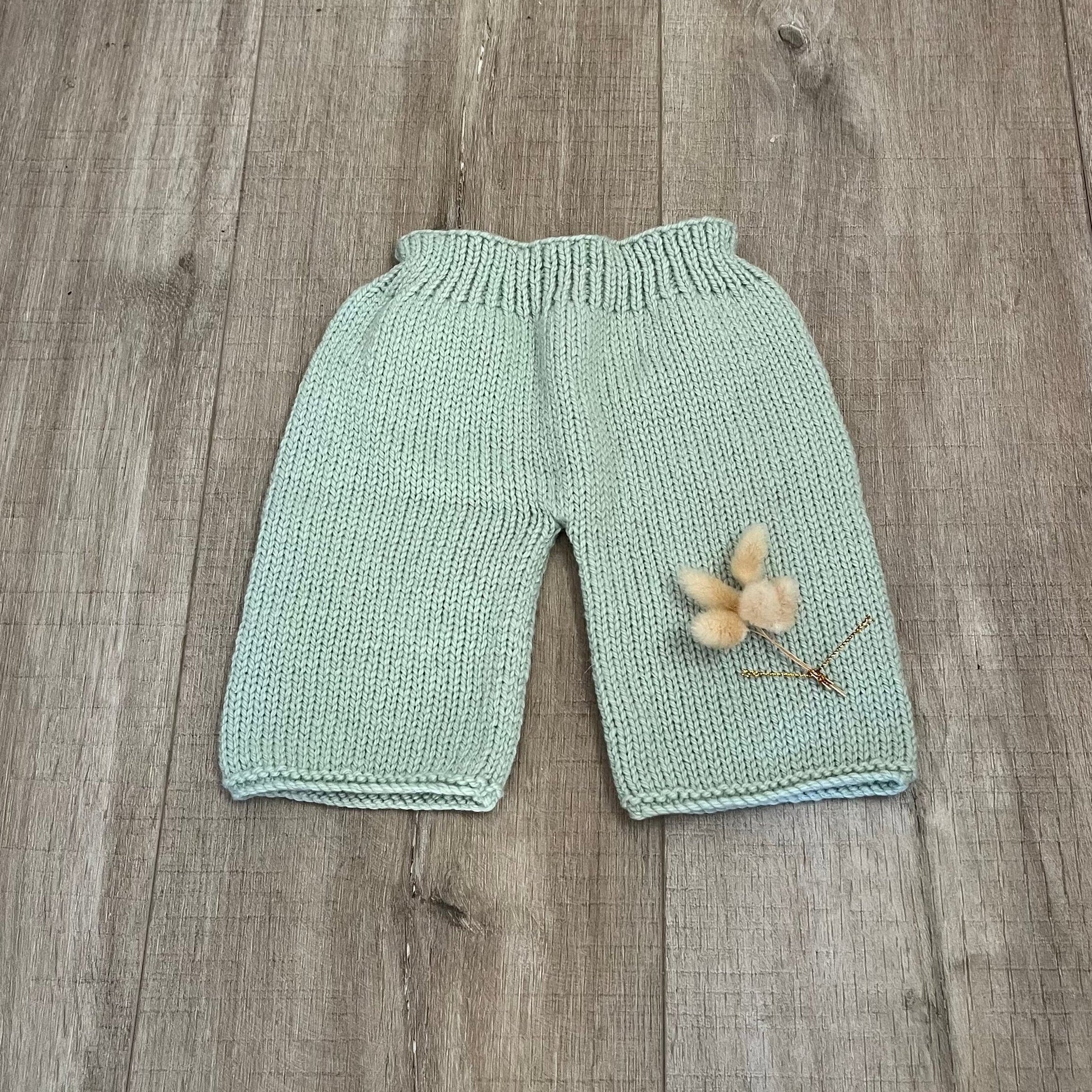 A pair of light mint green hand-knit baby pants with a ribbed waistband, laid flat on a gray-brown wooden floor with a small dried flower prop on the leg.