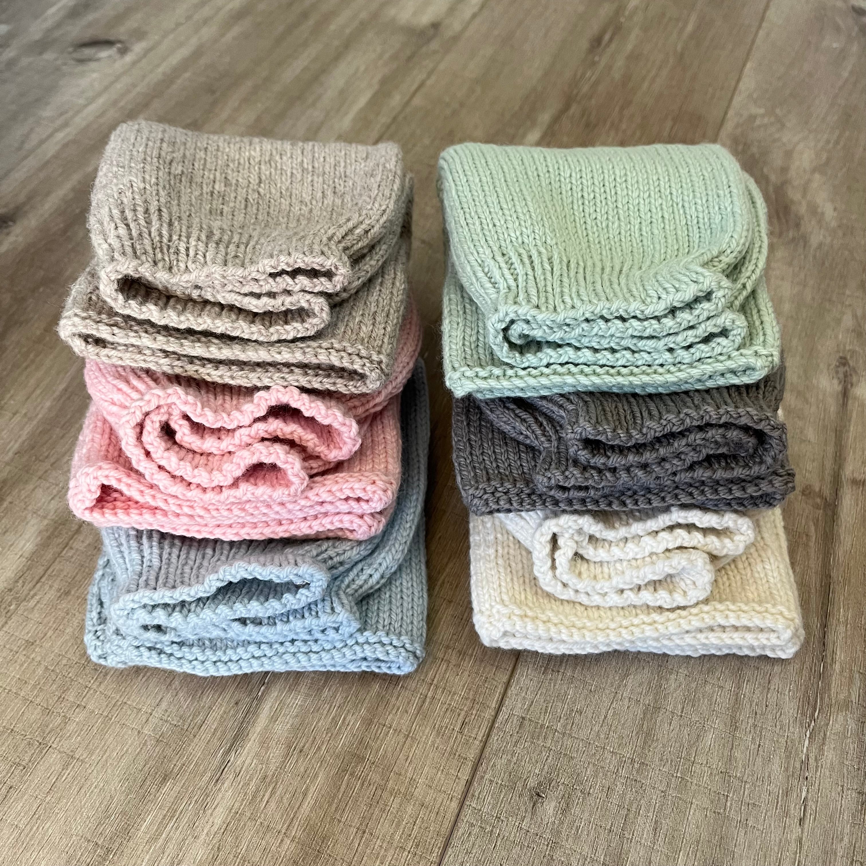 six folded ribbed knit baby pants in neutral and pastel colors, arranged in two stacks on a wooden floor. The colors include taupe, sage green, dusty pink, light blue, charcoal gray, and cream.