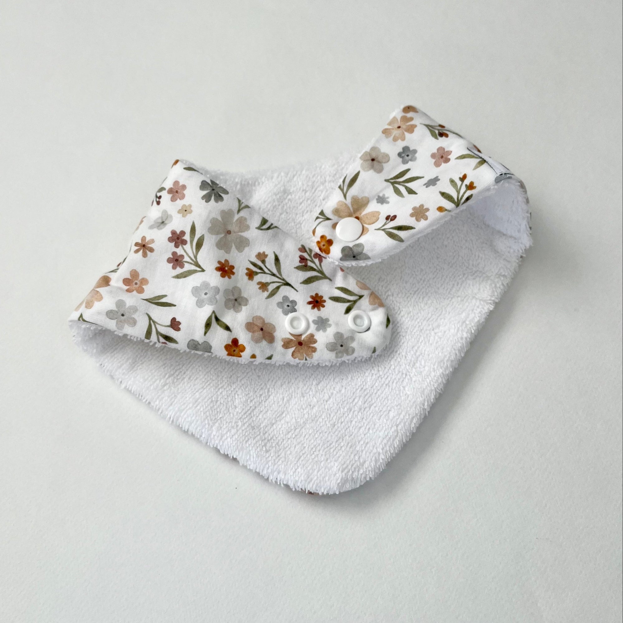 A reusable baby bib with a white terry cloth body and a patterned fabric flap featuring small flowers and leaves in muted brown, gray, and olive tones, shown on a light gray background.