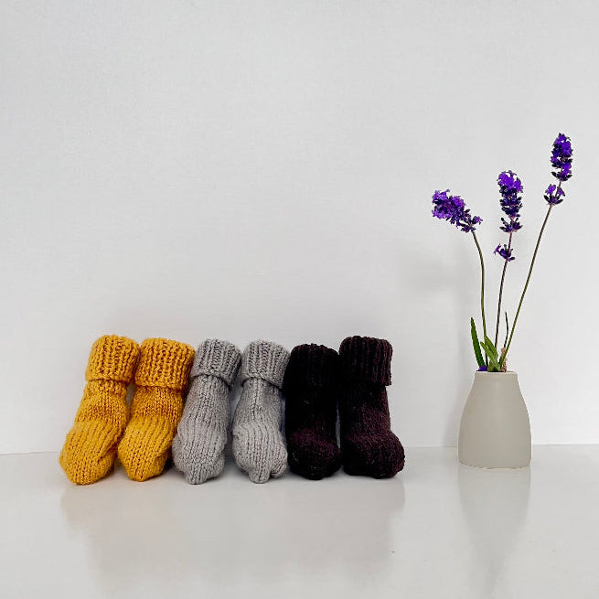 Knitted socks in mustard, mushroom, and chocolate on a white surface with a vase of lavender.