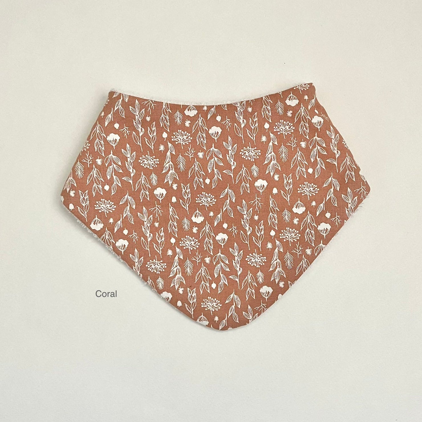 A coral-colored triangular bandana bib with a white floral and leaf pattern, laid flat on a light off-white background with the word 'Coral' printed near the bottom left.