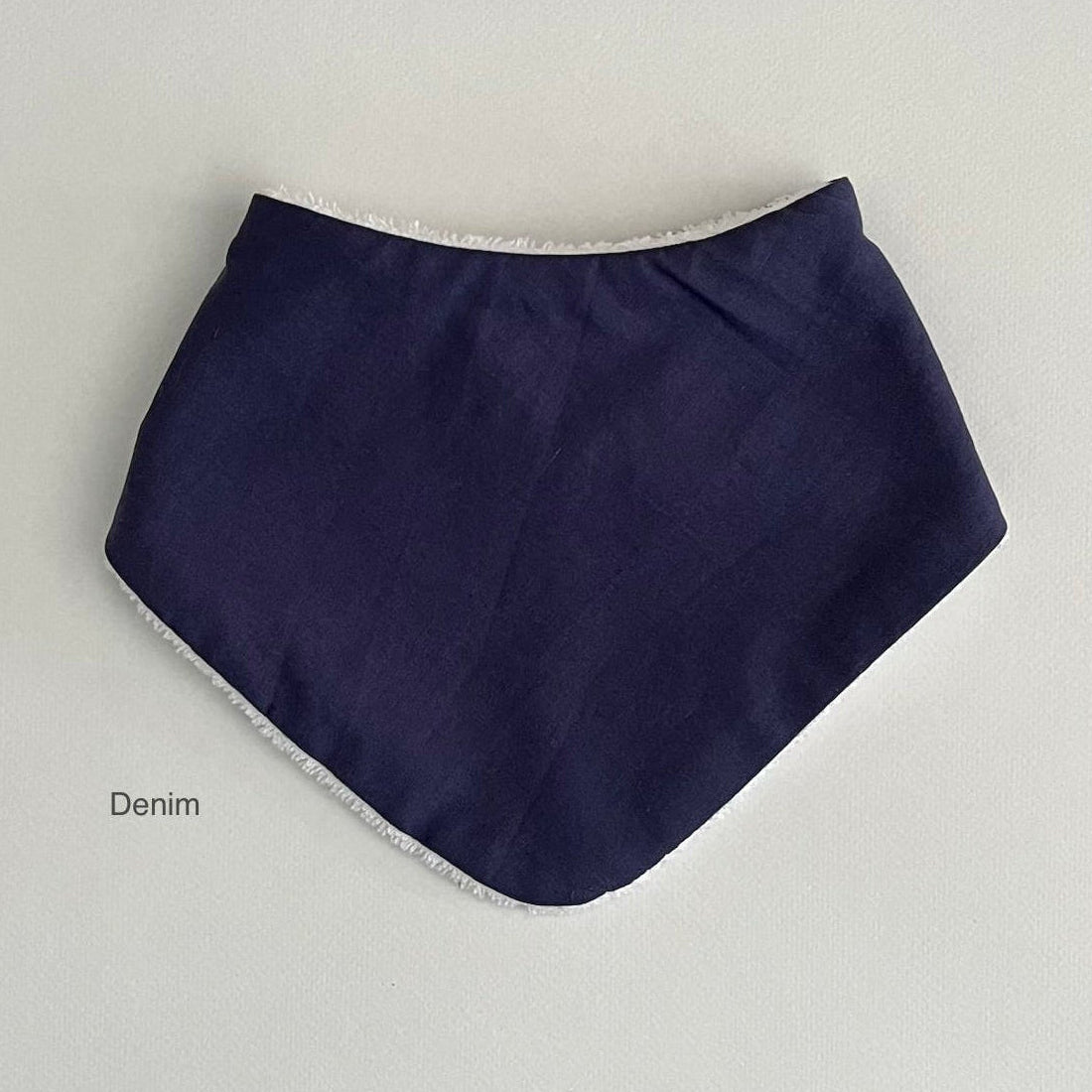 A navy blue triangular bib or bandana-style baby accessory laid flat on a light gray background, with a white frayed trim along the edges. The word 'Denim' appears in small text near the lower left.