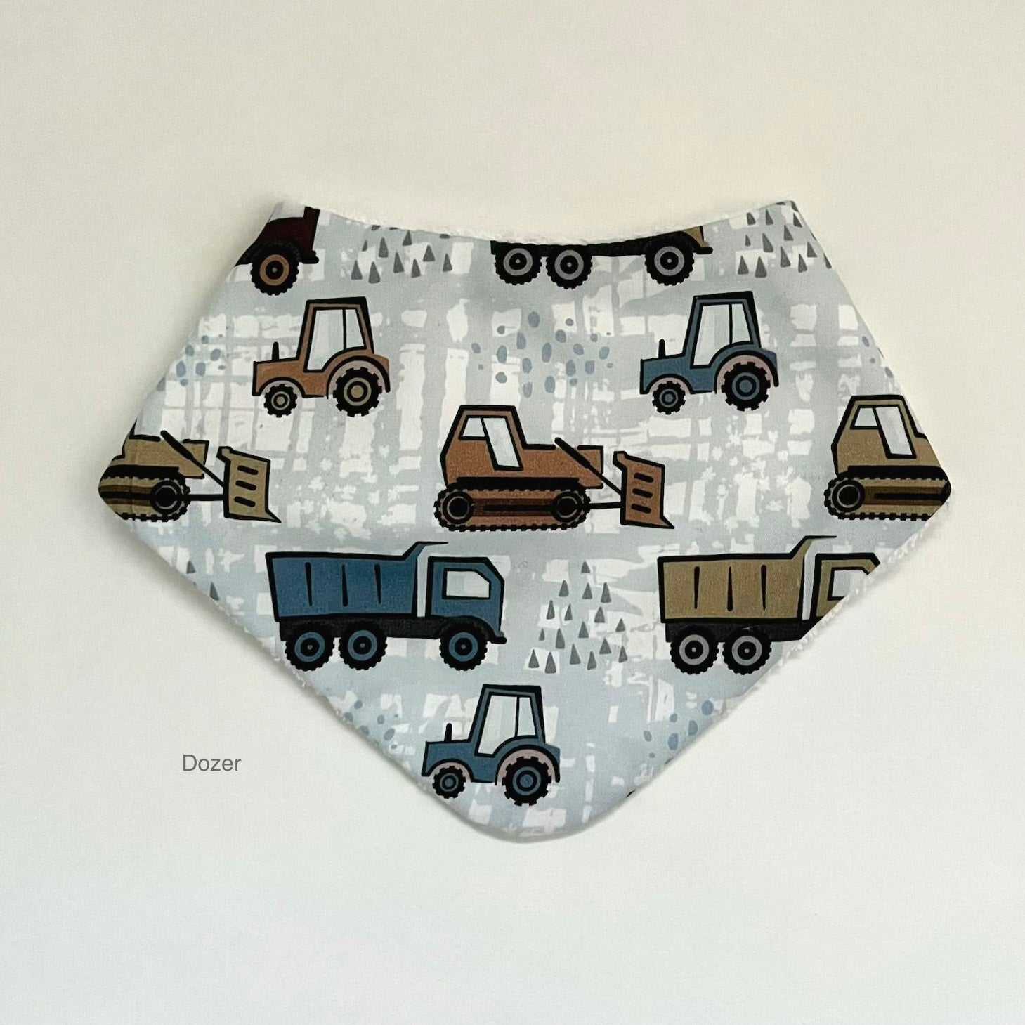 A triangular bandana bib with a construction vehicle pattern, featuring dump trucks, bulldozers, and tractors on a light gray background, displayed against a plain off-white wall.
