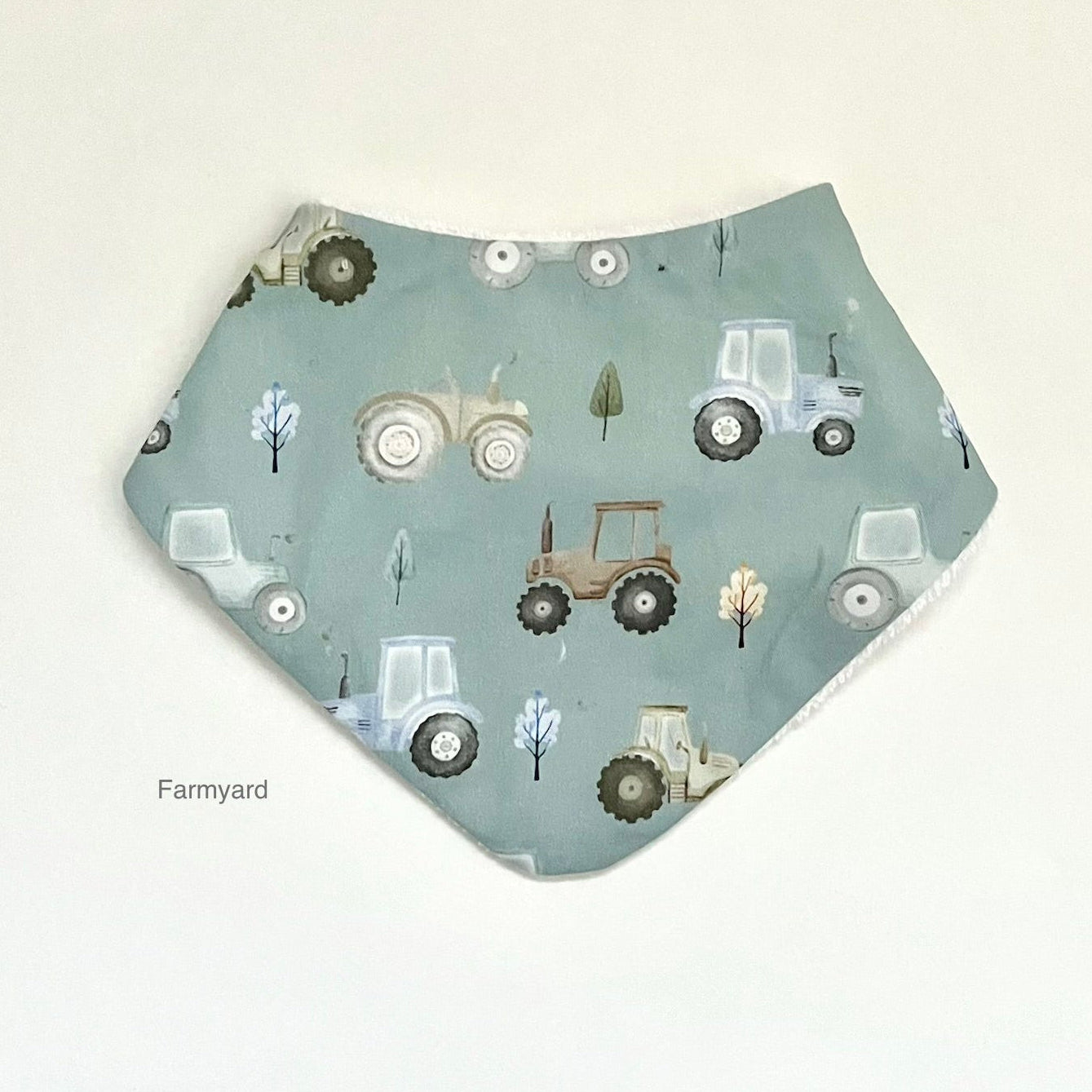 A triangular baby bandana bib with a sage green background and a pattern of various farm tractors and trees, shown on a plain off-white background with a 'Farmyard' watermark.
