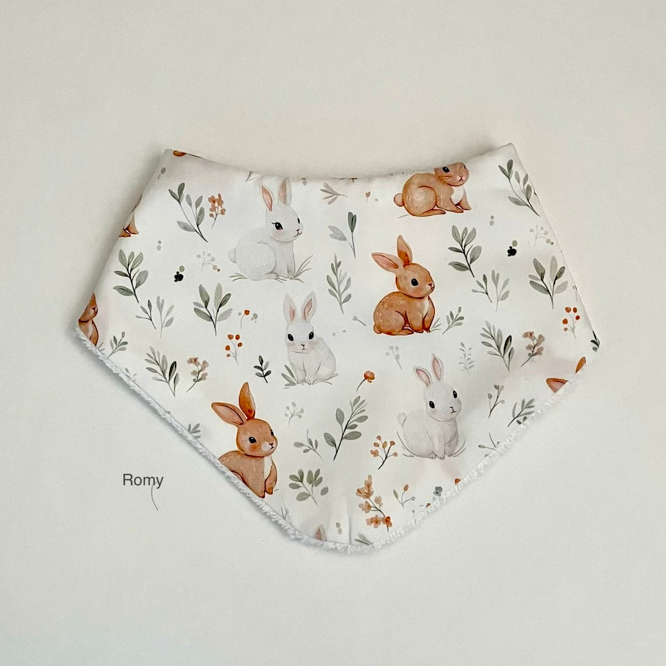 A triangular bandana baby bib with a white background and a printed pattern of brown and white rabbits, leaves, and small flowers, laid flat on a light beige surface.