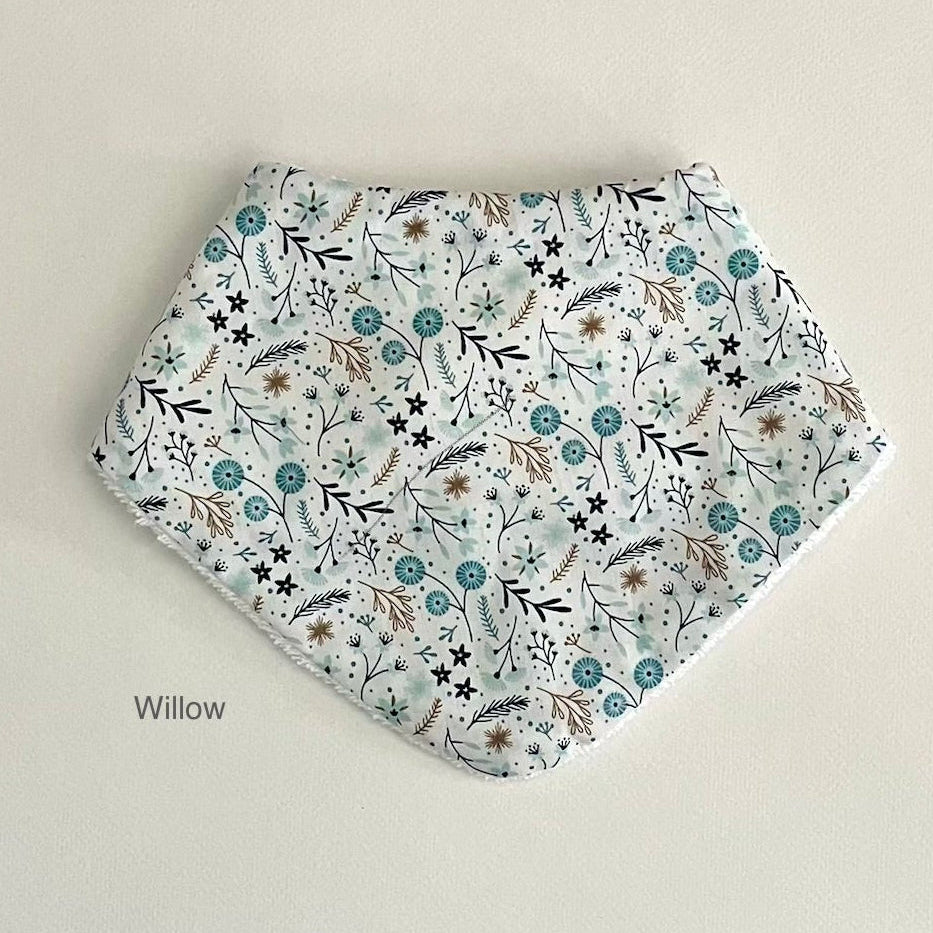 A triangular bandana bib with a delicate floral and botanical print in teal, brown, and black on a white background, displayed flat against a light beige background with the text 'Willow' printed below.