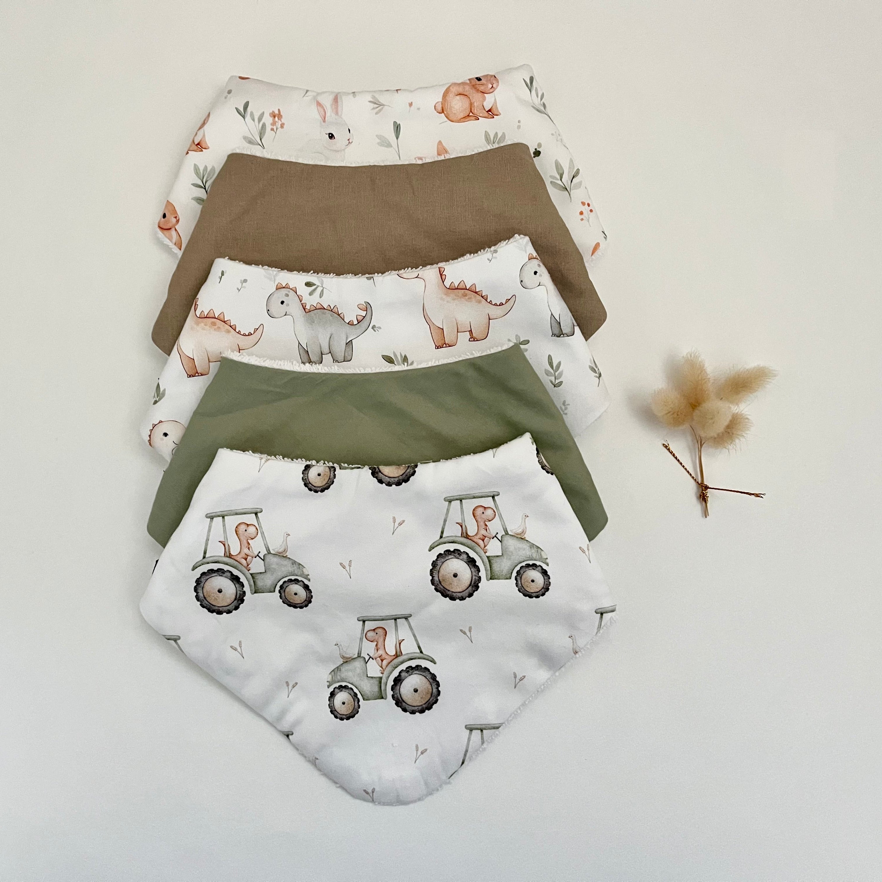 Five reusable baby bibs stacked on a white background, featuring tractor, dinosaur, and elephant prints in soft earthy tones, with a small dried flower prop on the right.