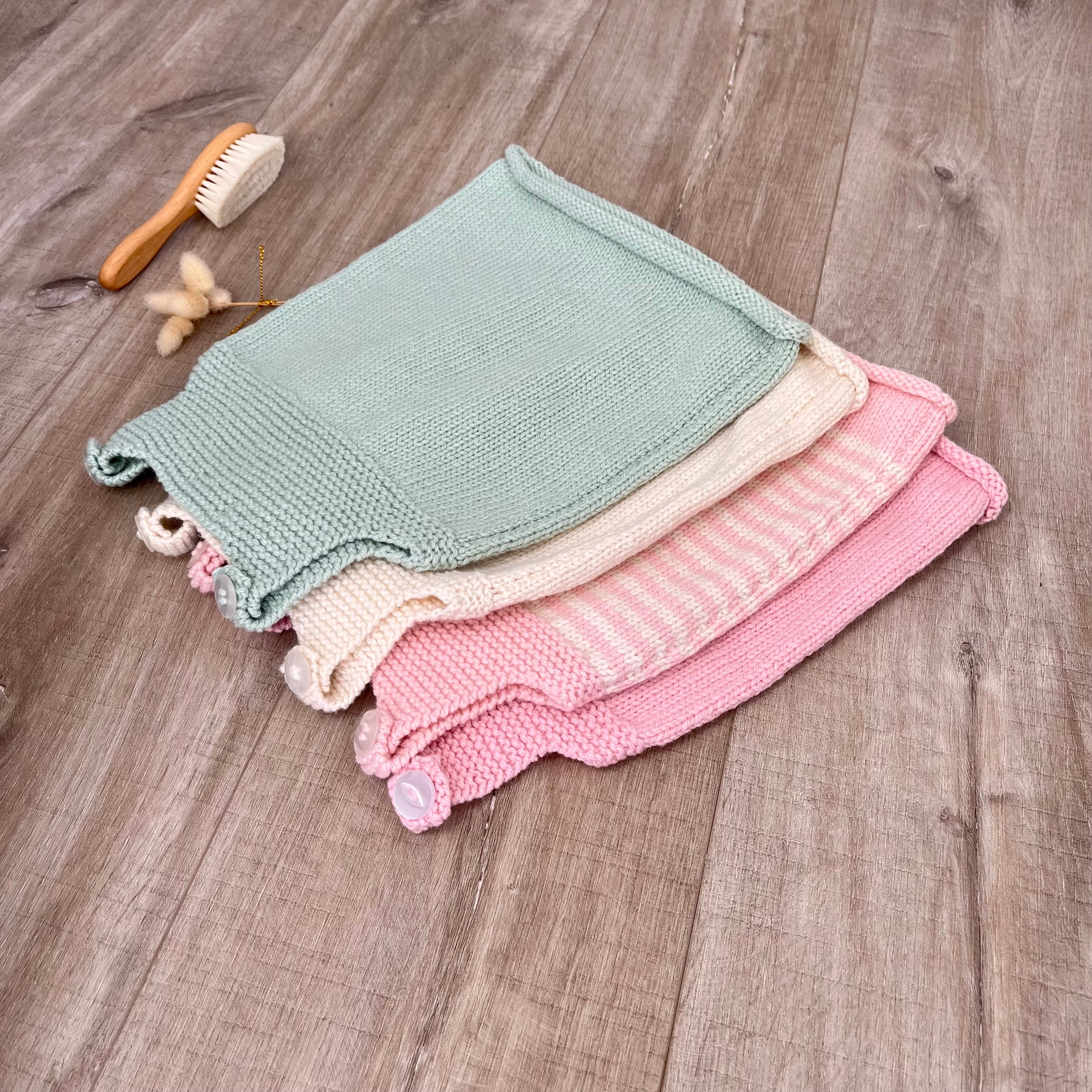 Three folded knitted baby dresses in sage green, cream, and pink are stacked on a rustic wood floor, with a small wooden brush and dried flowers nearby.