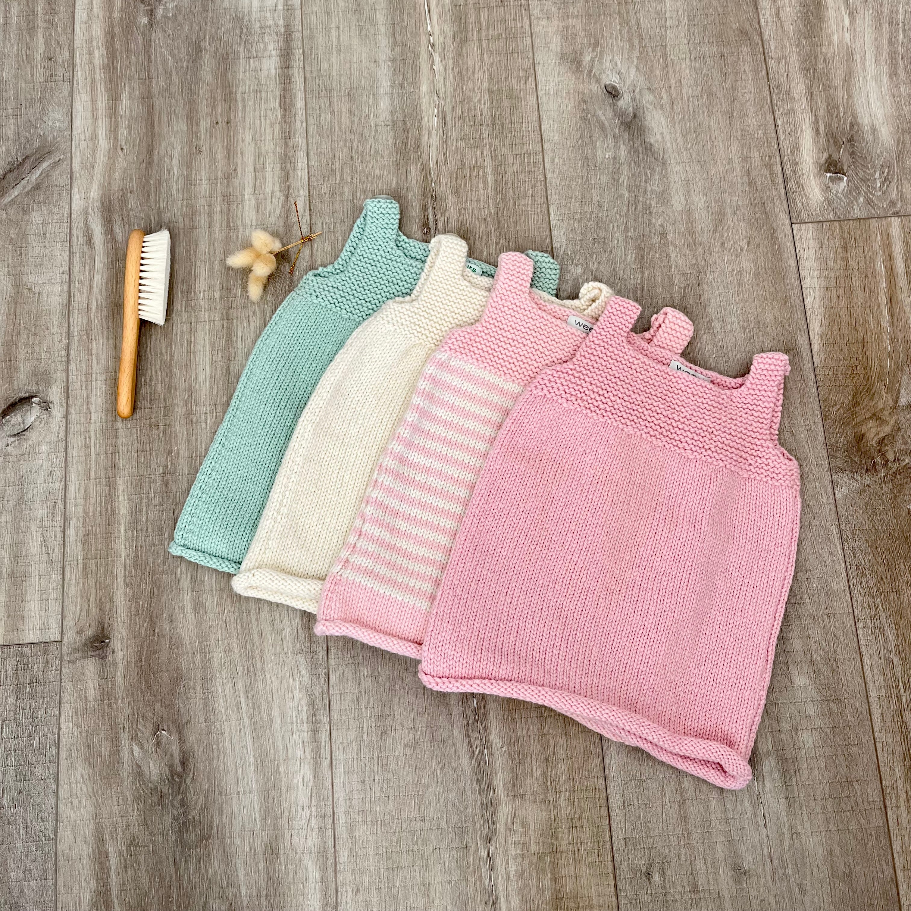 Four knitted baby dresses in pastel colors (mint green, cream, pink, and pink-and-white stripes) arranged on a rustic wood plank floor next to a wooden baby toothbrush and dried flowers.