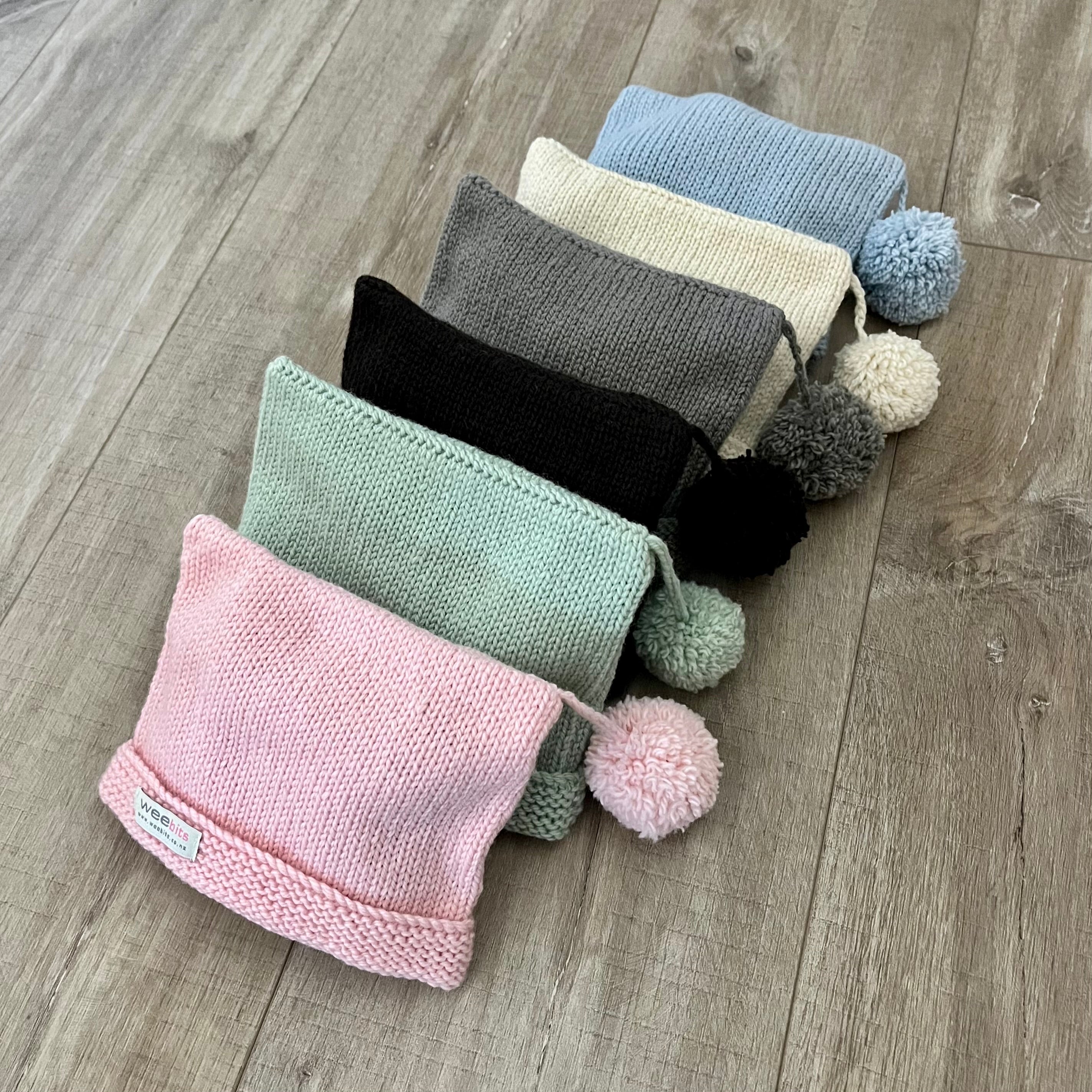 Six knitted pouches or small hats arranged diagonally on a wood floor, each topped with a pom-pom. The hats come in pastel pink, mint green, black, charcoal gray, cream, and light blue, with the front pink one showing a small white brand tag.