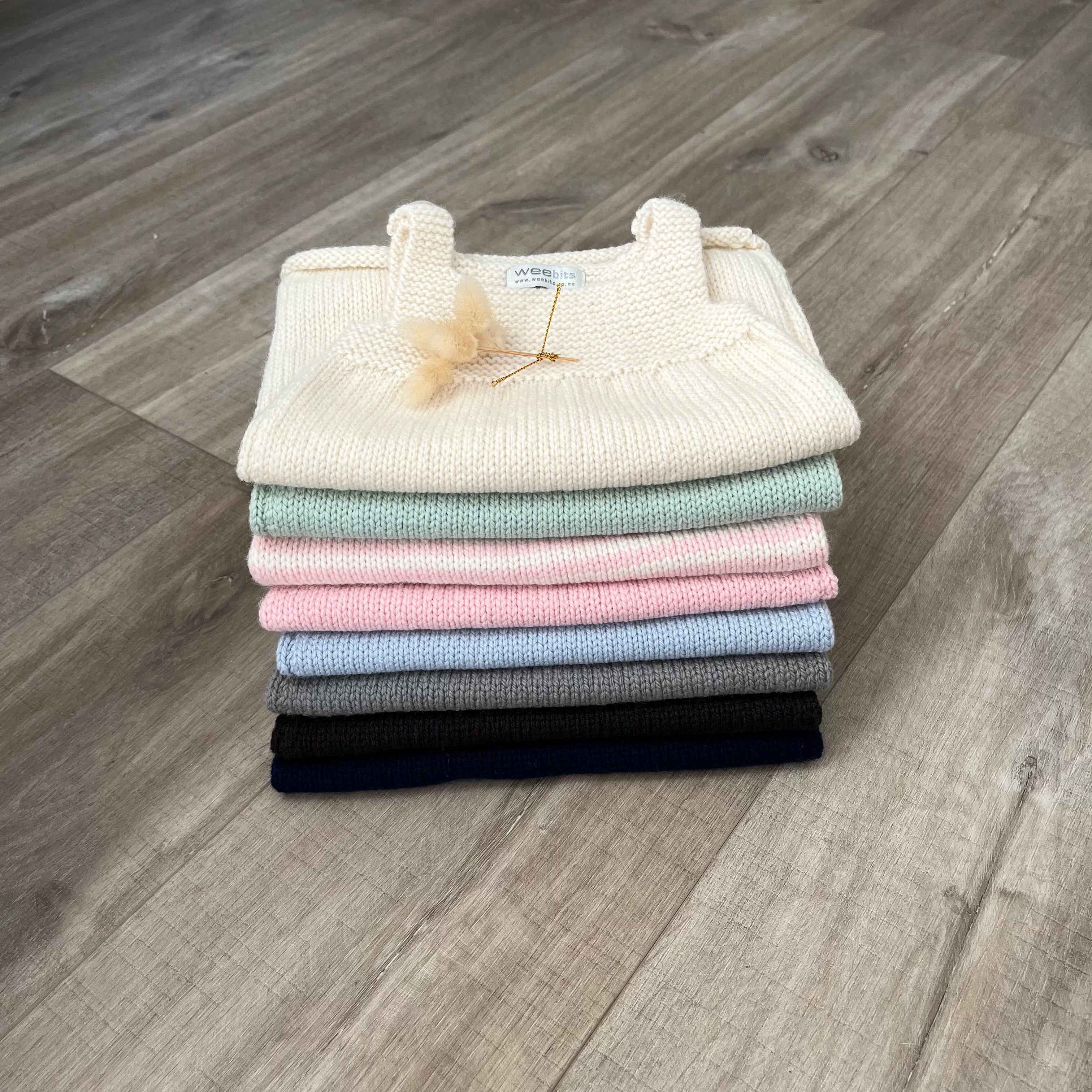 A stack of folded knit dresses in cream, mint, light blue, pink, gray, brown, and navy, arranged on a gray wood-look floor. A small decorative flower is tucked into the top cream sweater.