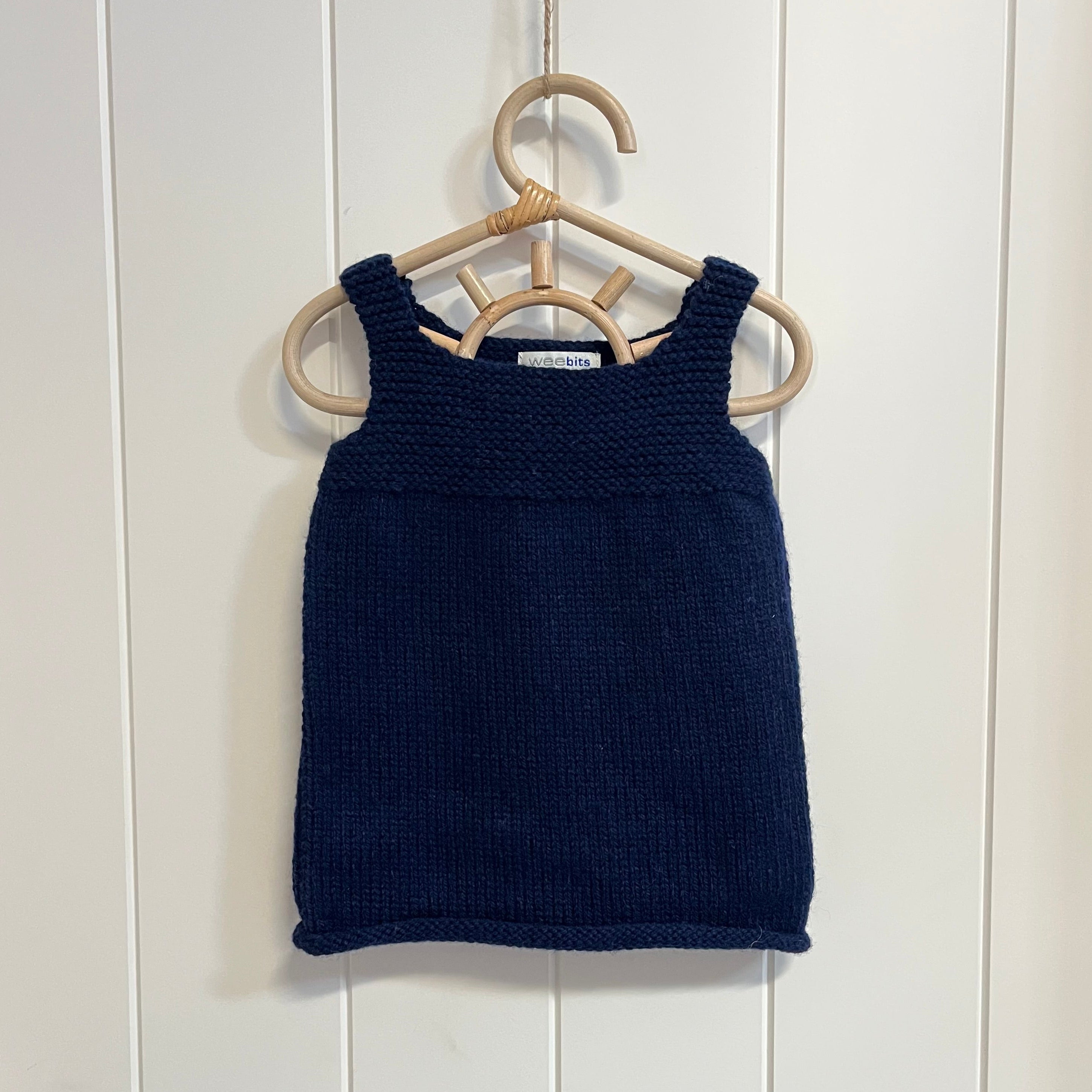 A navy hand-knit sleeveless dress hanging on a light wood hanger against a white paneled wall.