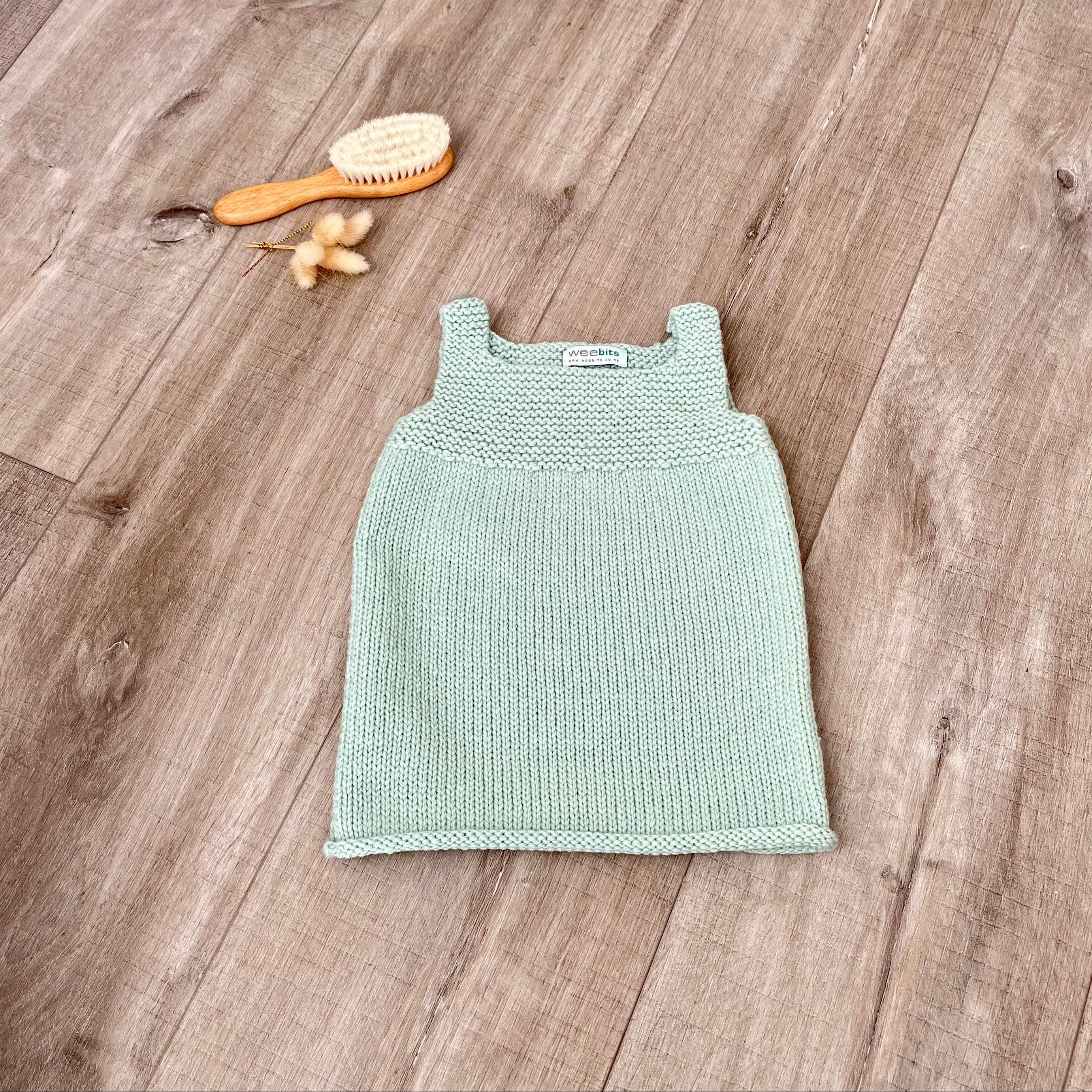 A mint knit sleeveless baby dress laid flat on a light brown wooden surface, styled with a small orange-handled hairbrush and dried flowers near the top left.