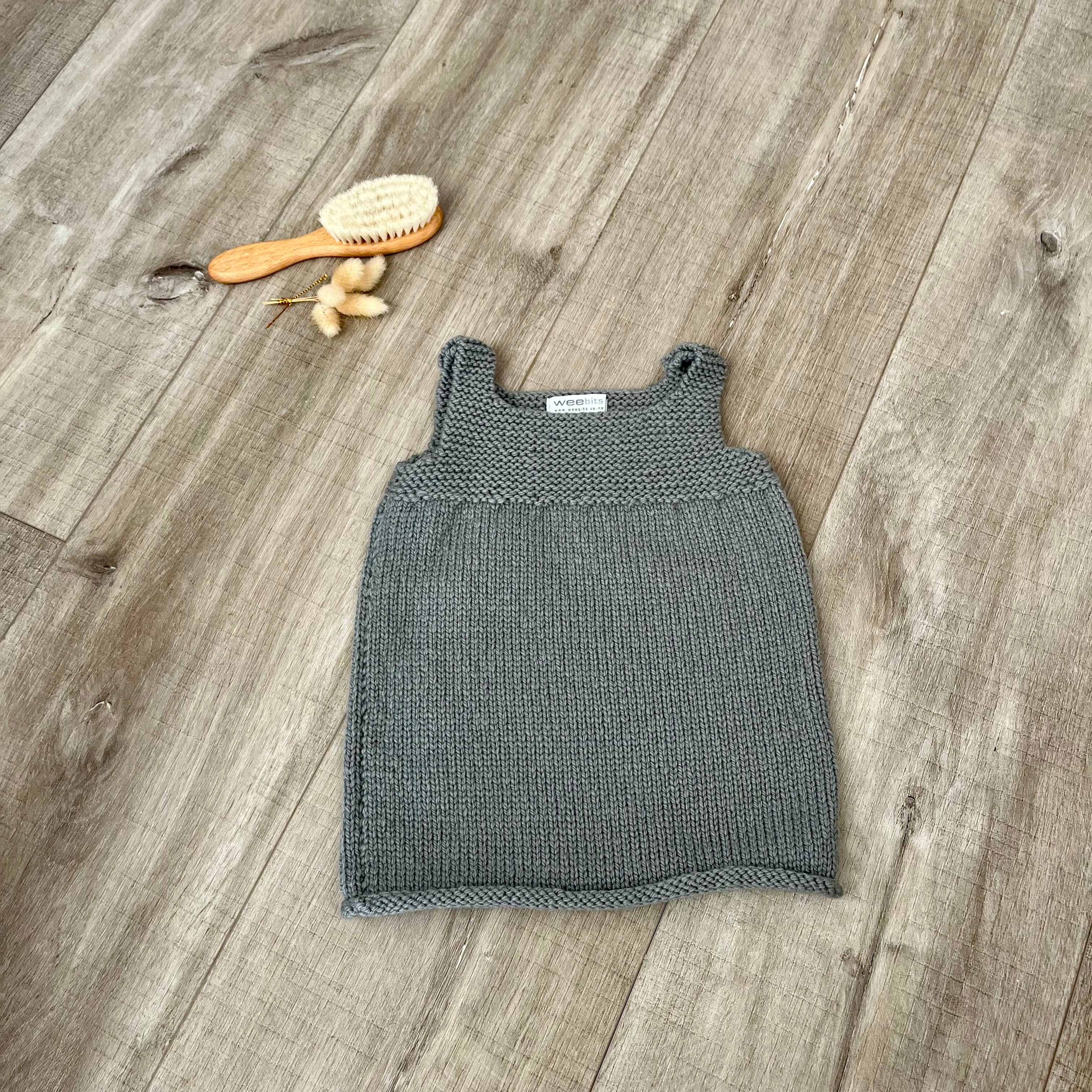 A mushroom knit sleeveless baby dress laid flat on a light brown wooden surface, styled with a small orange-handled hairbrush and dried flowers near the top left.