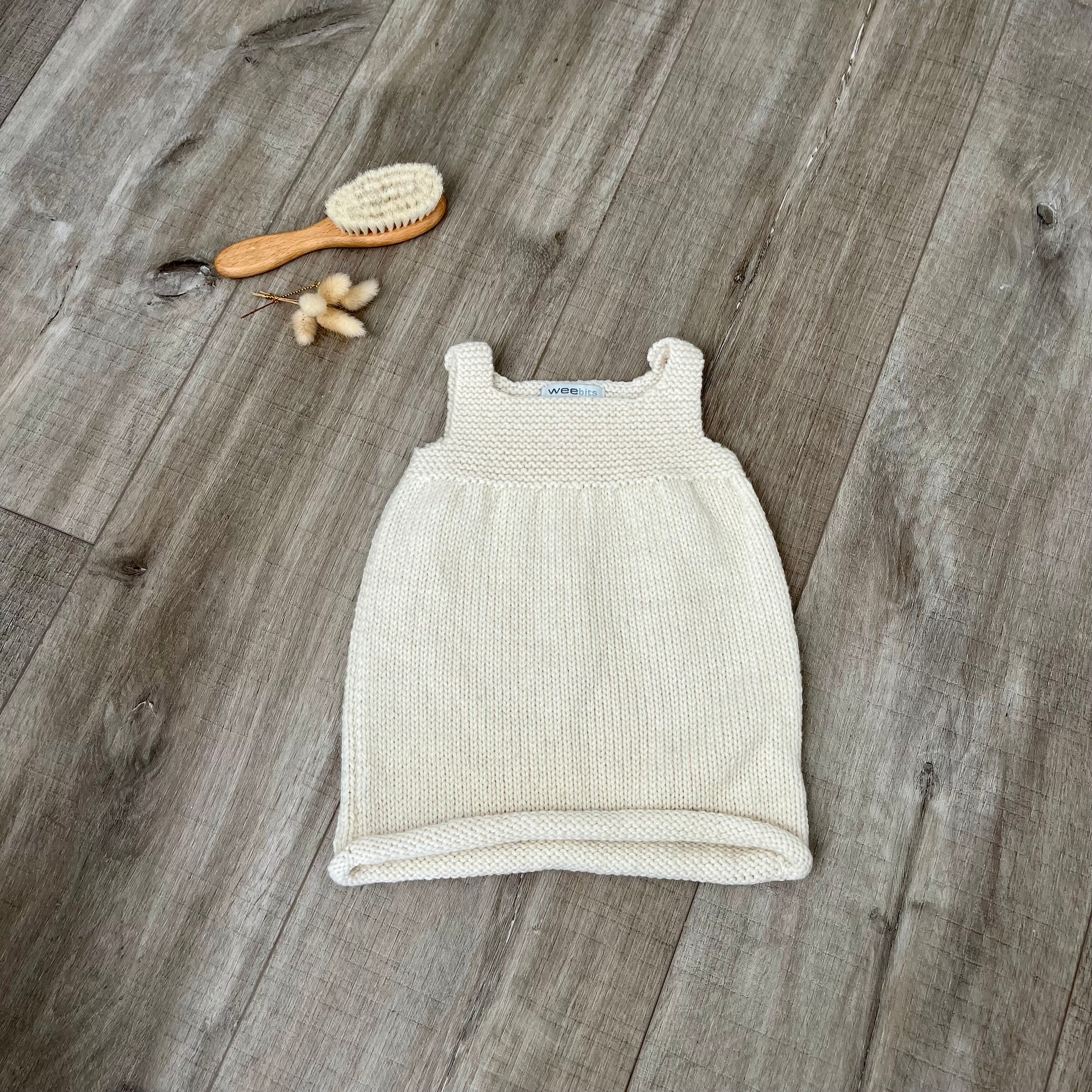 A natural knit sleeveless baby dress laid flat on a light brown wooden surface, styled with a small orange-handled hairbrush and dried flowers near the top left.