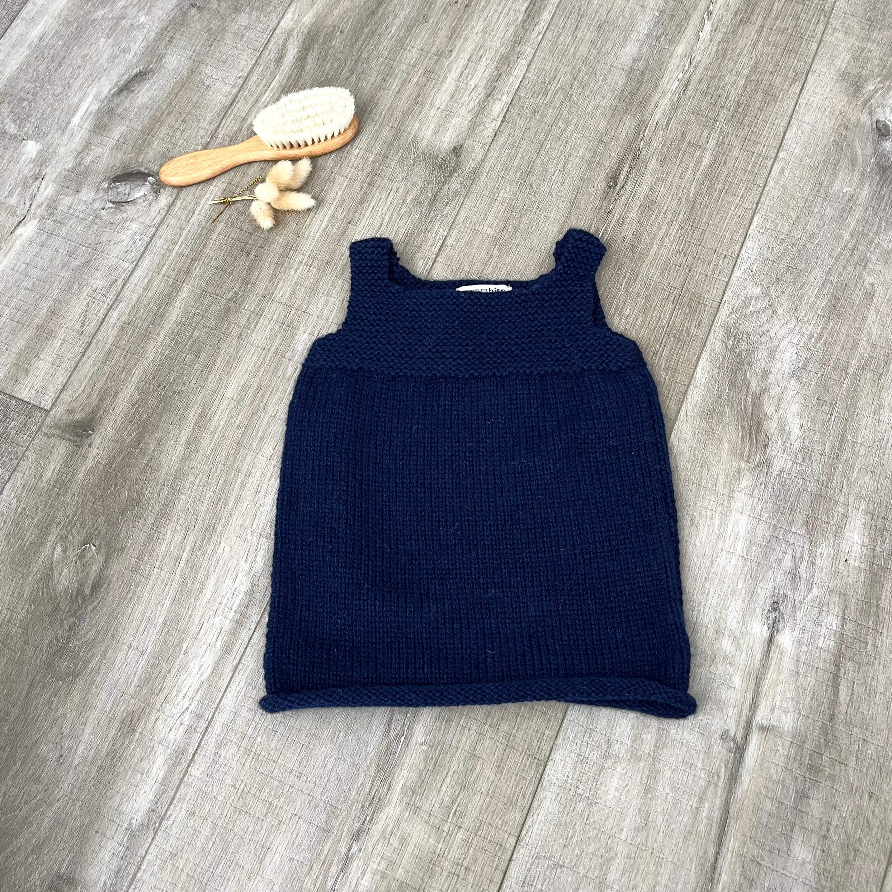 A navy knit sleeveless baby dress laid flat on a light brown wooden surface, styled with a small orange-handled hairbrush and dried flowers near the top left.