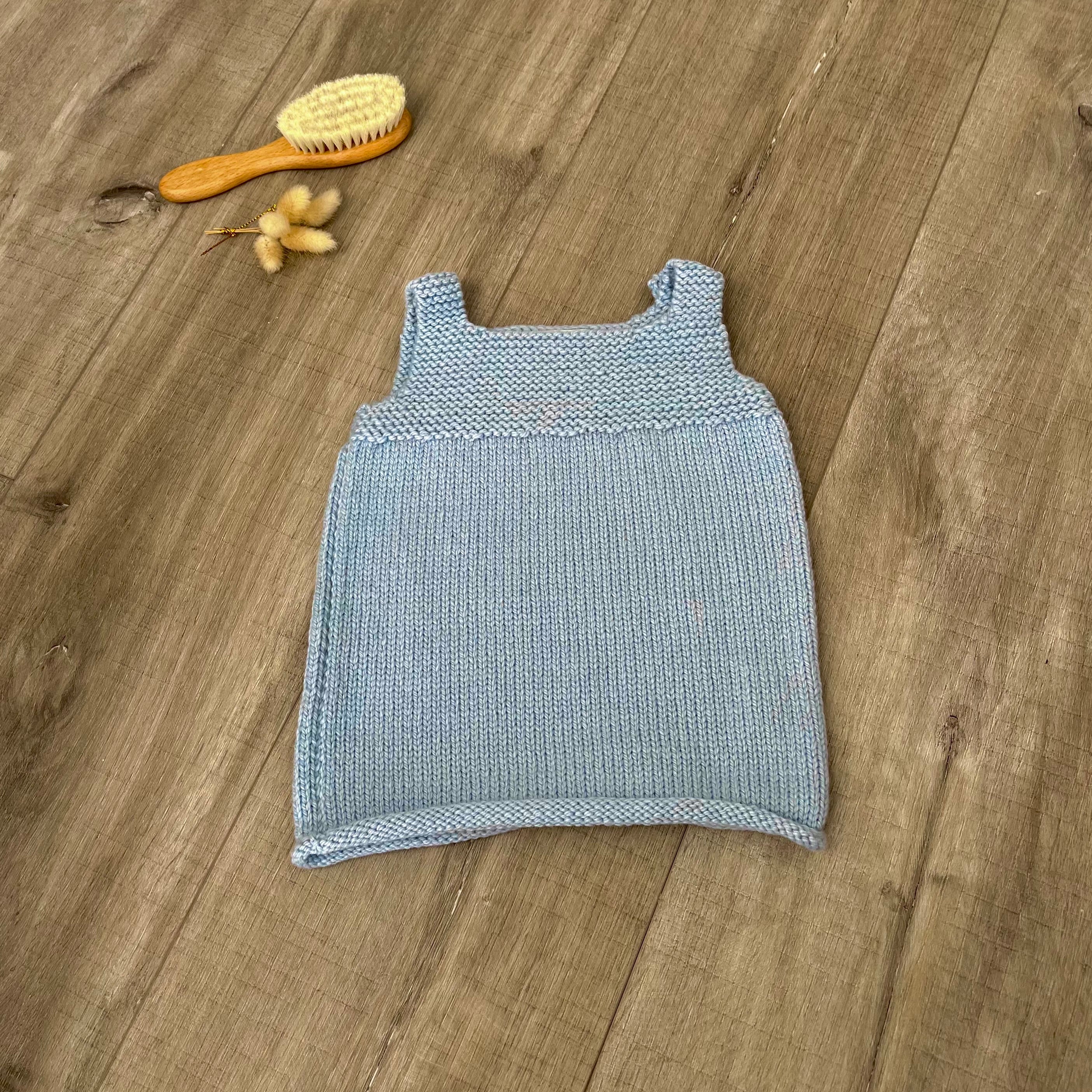 A blue knit sleeveless baby dress laid flat on a light brown wooden surface, styled with a small orange-handled hairbrush and dried flowers near the top left.