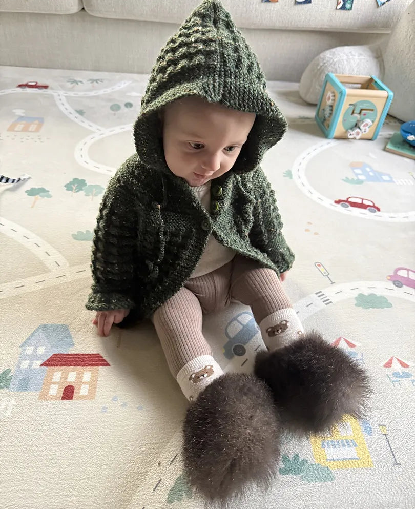 A baby sitting on a printed play mat wearing a dark green textured knit hooded jacket, beige knit pants, and fluffy dark brown possum booties