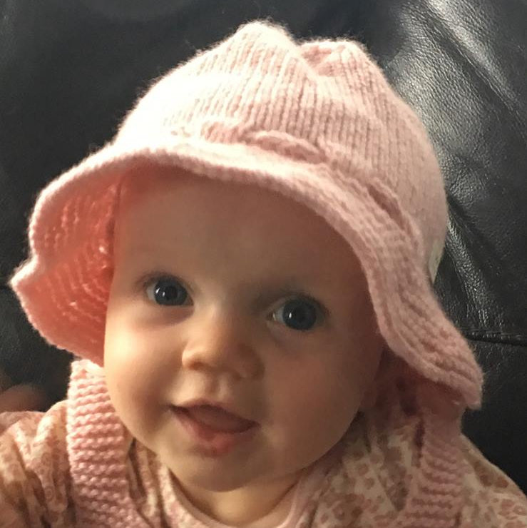 A close-up of a baby wearing a light pink hand-knit bucket hat and matching pink knit outfit against a dark leather background.