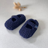 Pair of navy blue knitted baby shoes on a light gray surface with dried plants in the background.