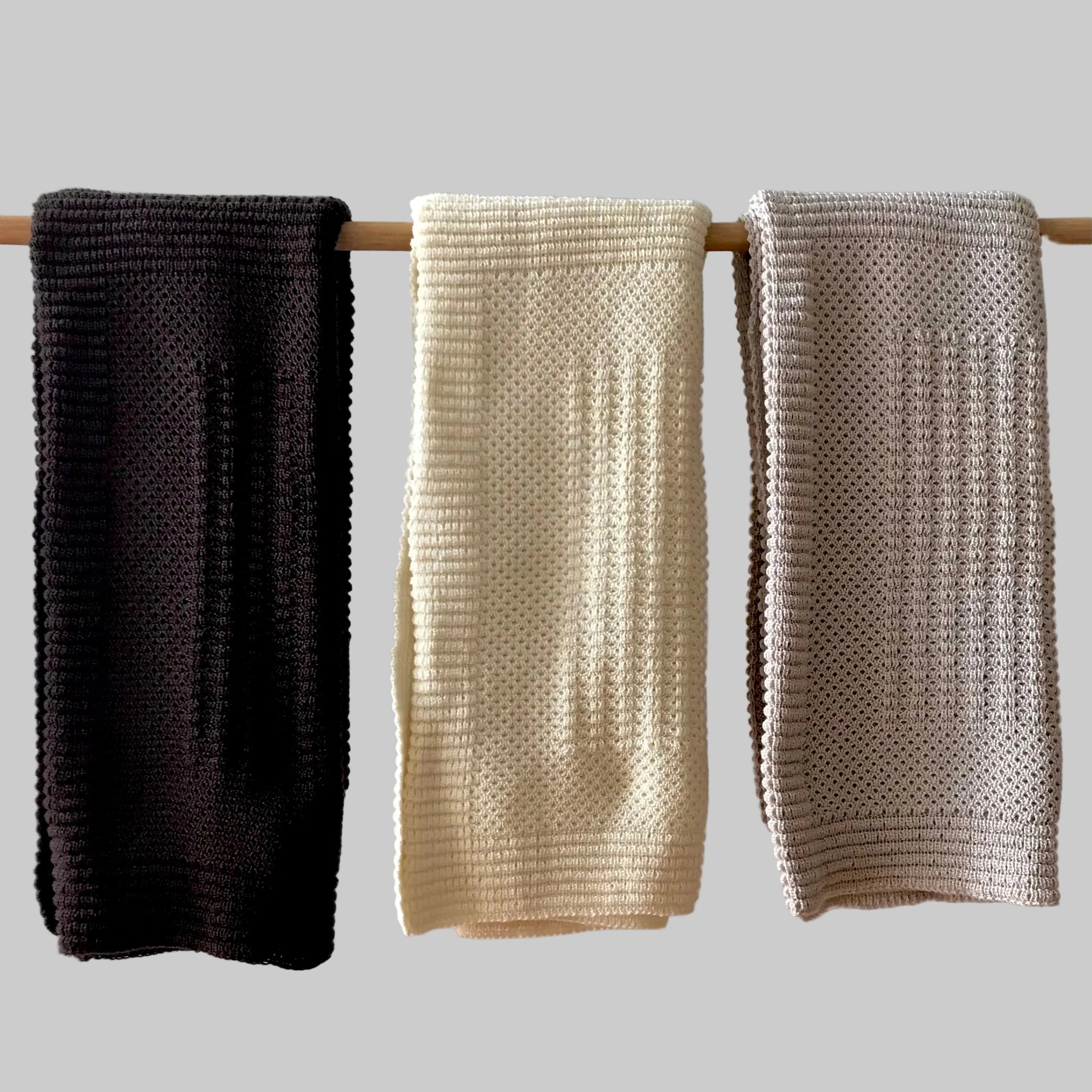 Three knitted cot blankets in brown, cream, and sandstone hanging on a rod against a neutral background.