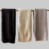 Three knitted cot blankets in brown, cream, and sandstone hanging on a rod against a neutral background.