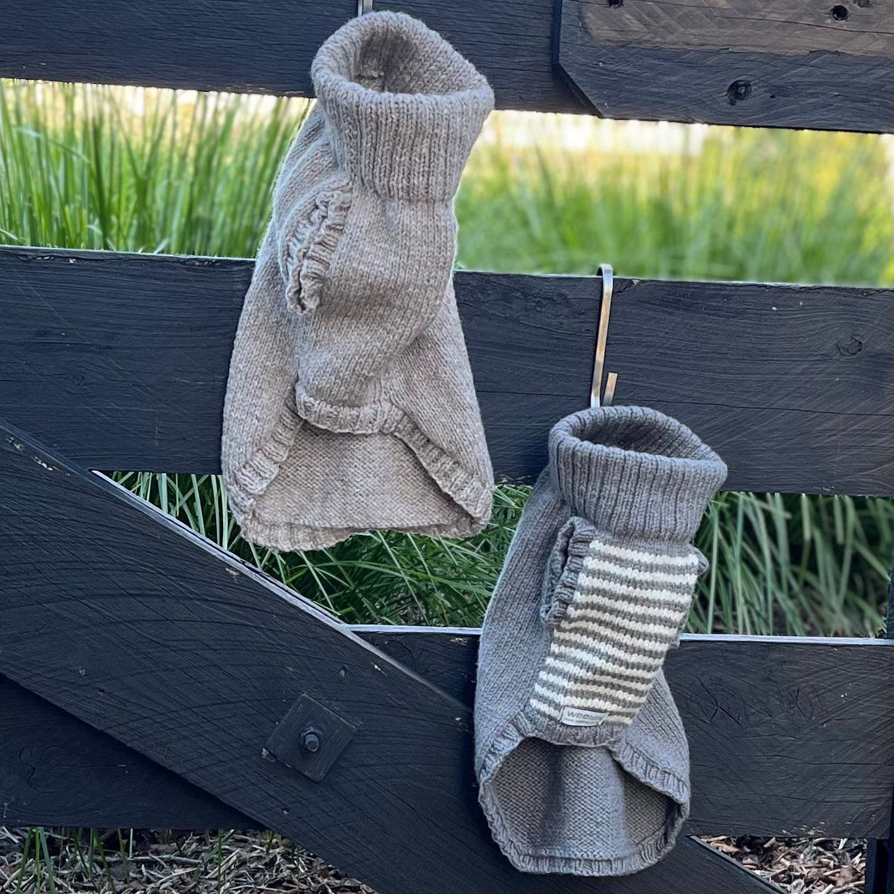 Two merino knitted dog jumpers hanging from hooks on a fence outside.