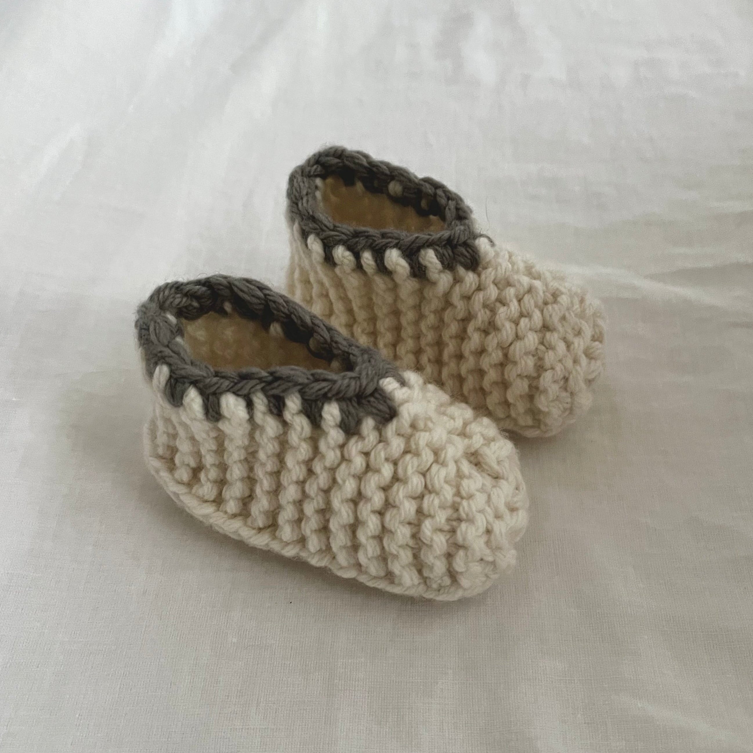 A pair of handmade, cream-colored merino slippers with dark gray trim, displayed on a soft, off-white fabric background.