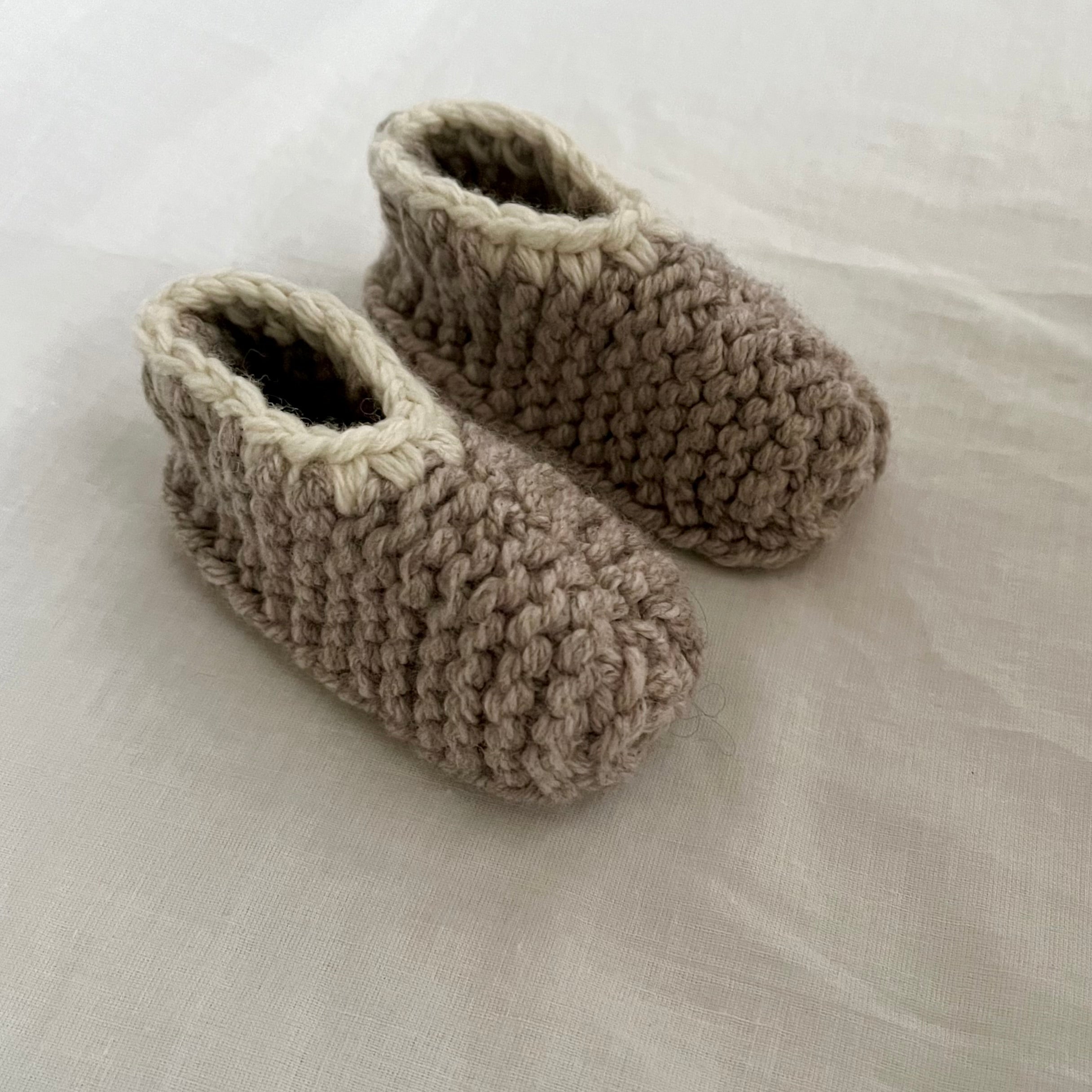 A pair of handmade, oatmeal-colored merino slippers with dark gray trim, displayed on a soft, off-white fabric background.