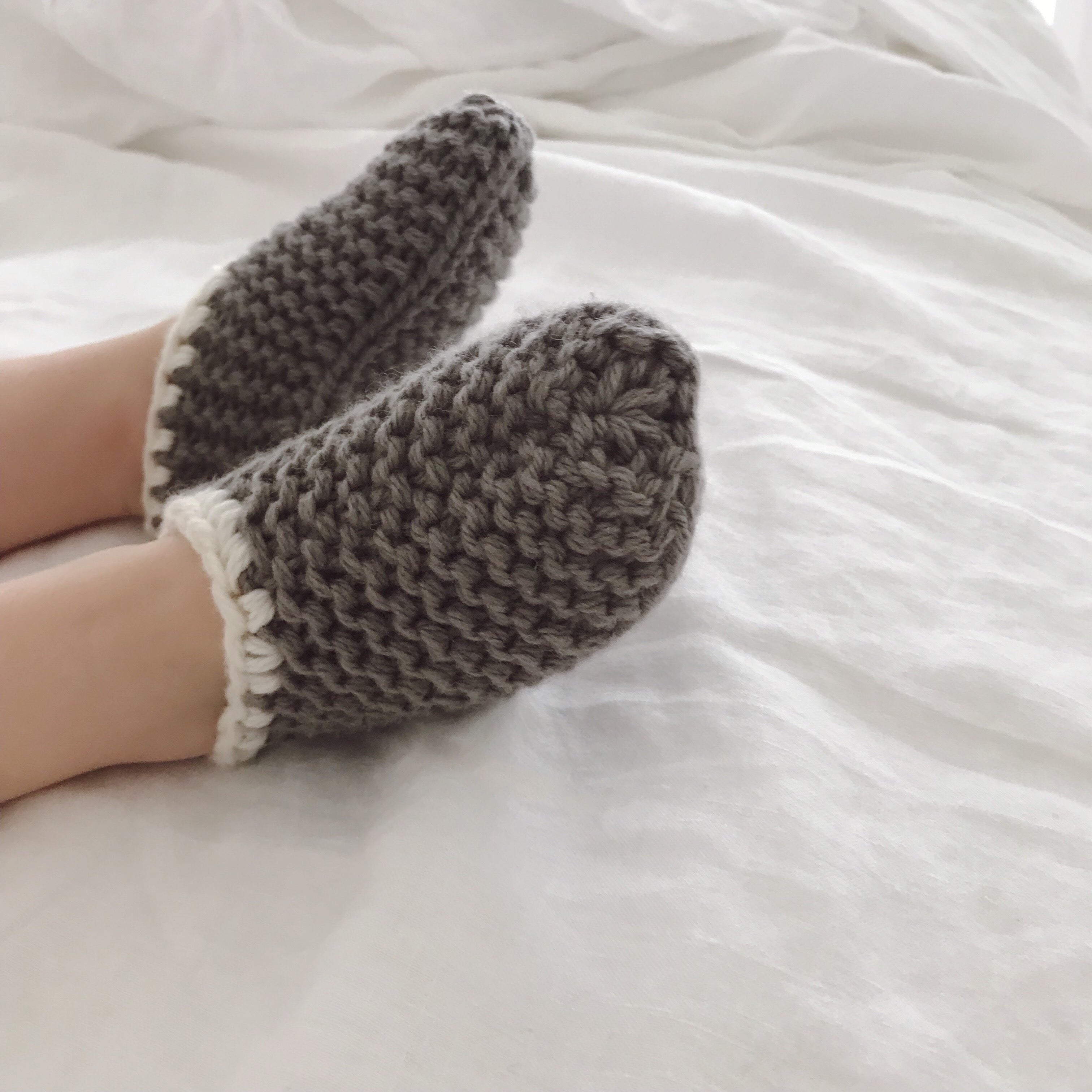 A pair of chunky hand-knit gray slipper socks with white trim, shown being worn on feet resting on white bedding.