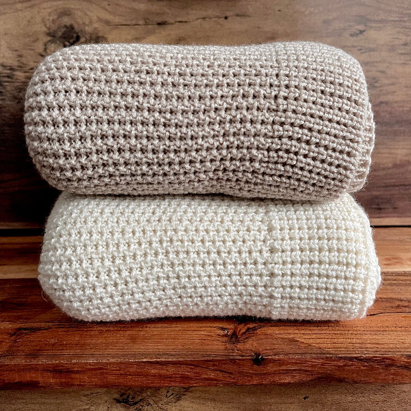 Two folded knitted blankets, one beige and one white, on a wooden surface.