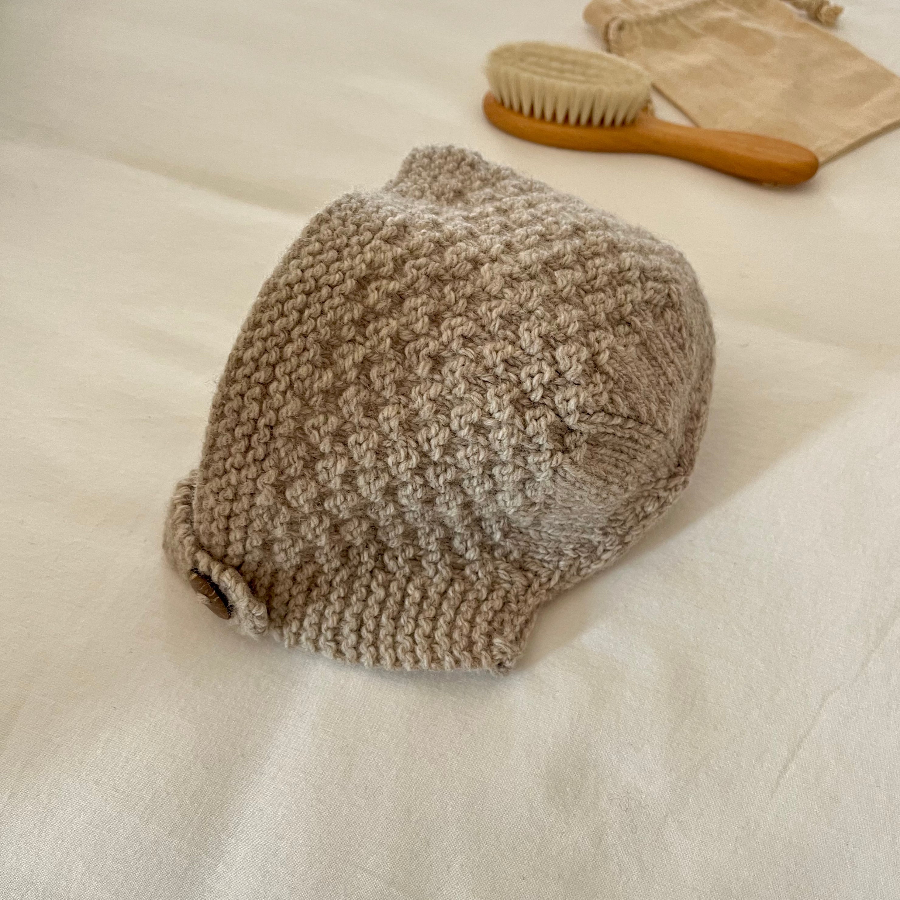 Knitted oatmeal bonnet on a white surface with a brush and cotton bag in the background