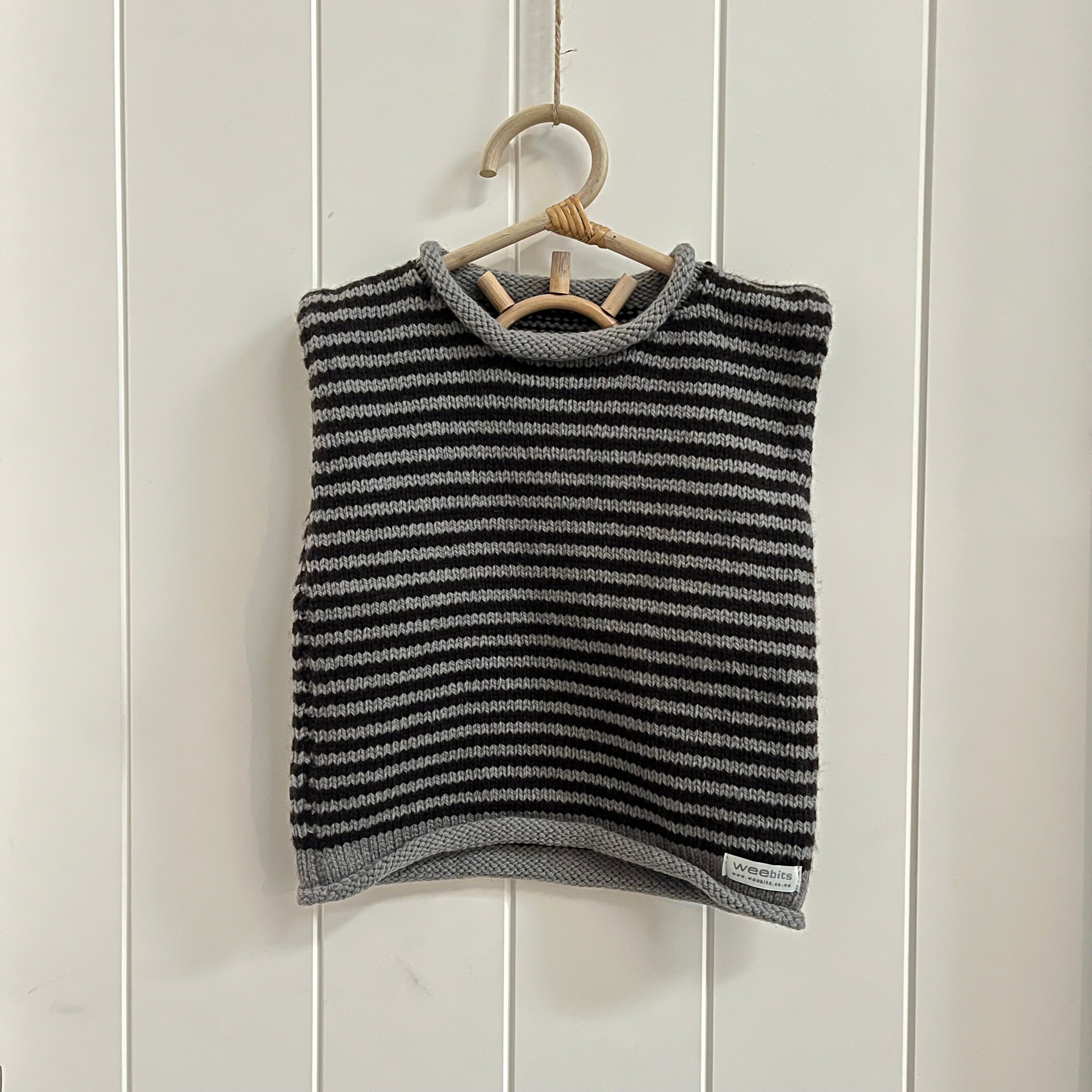 A dark gray and chocolate stripe knit sleeveless vest hanging on a light wood hanger against a white paneled wall.
