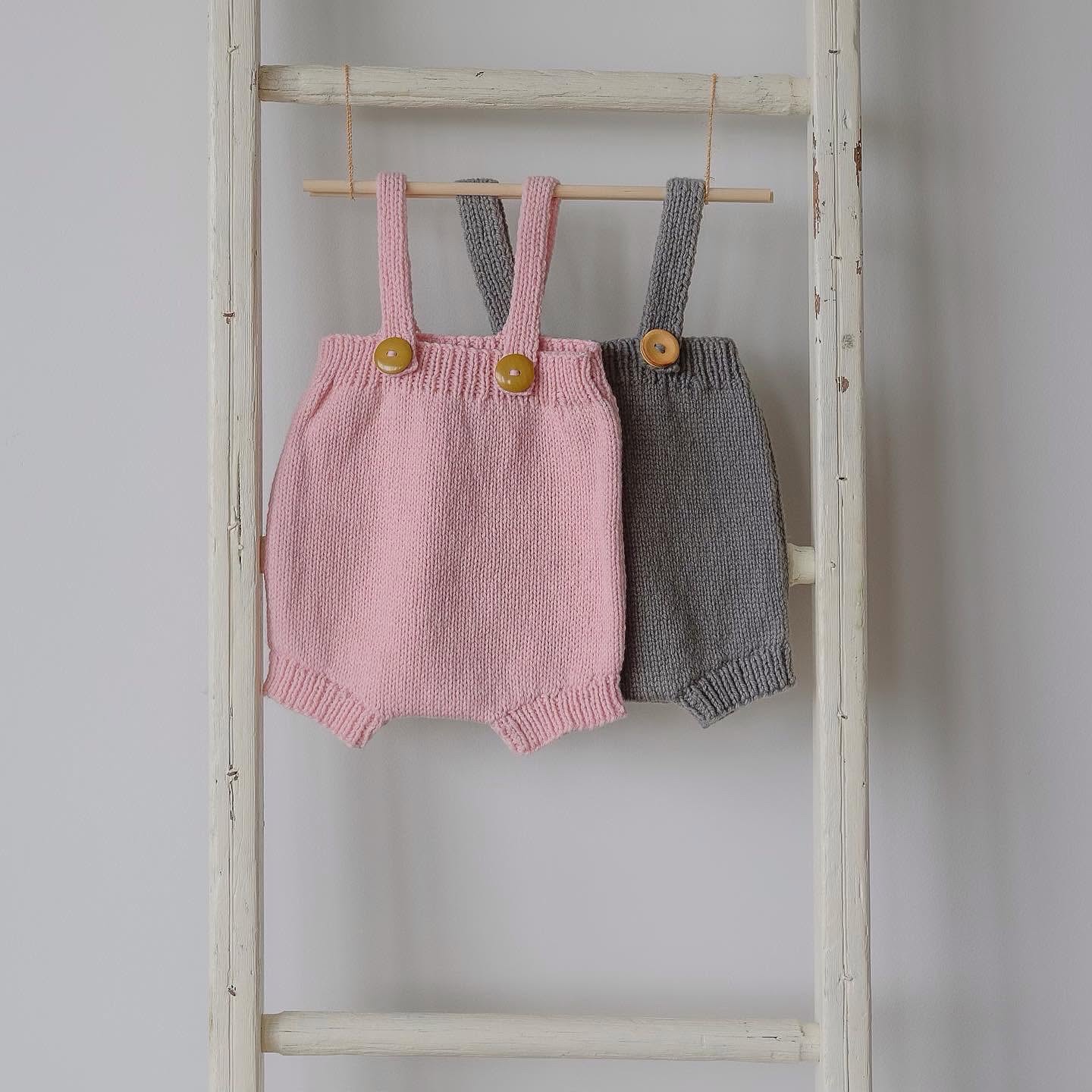 Two knitted baby romper shorts hanging from a rustic white wooden ladder. The front romper is light pink with two wooden buttons, and the one behind it is heather gray with a wooden button.