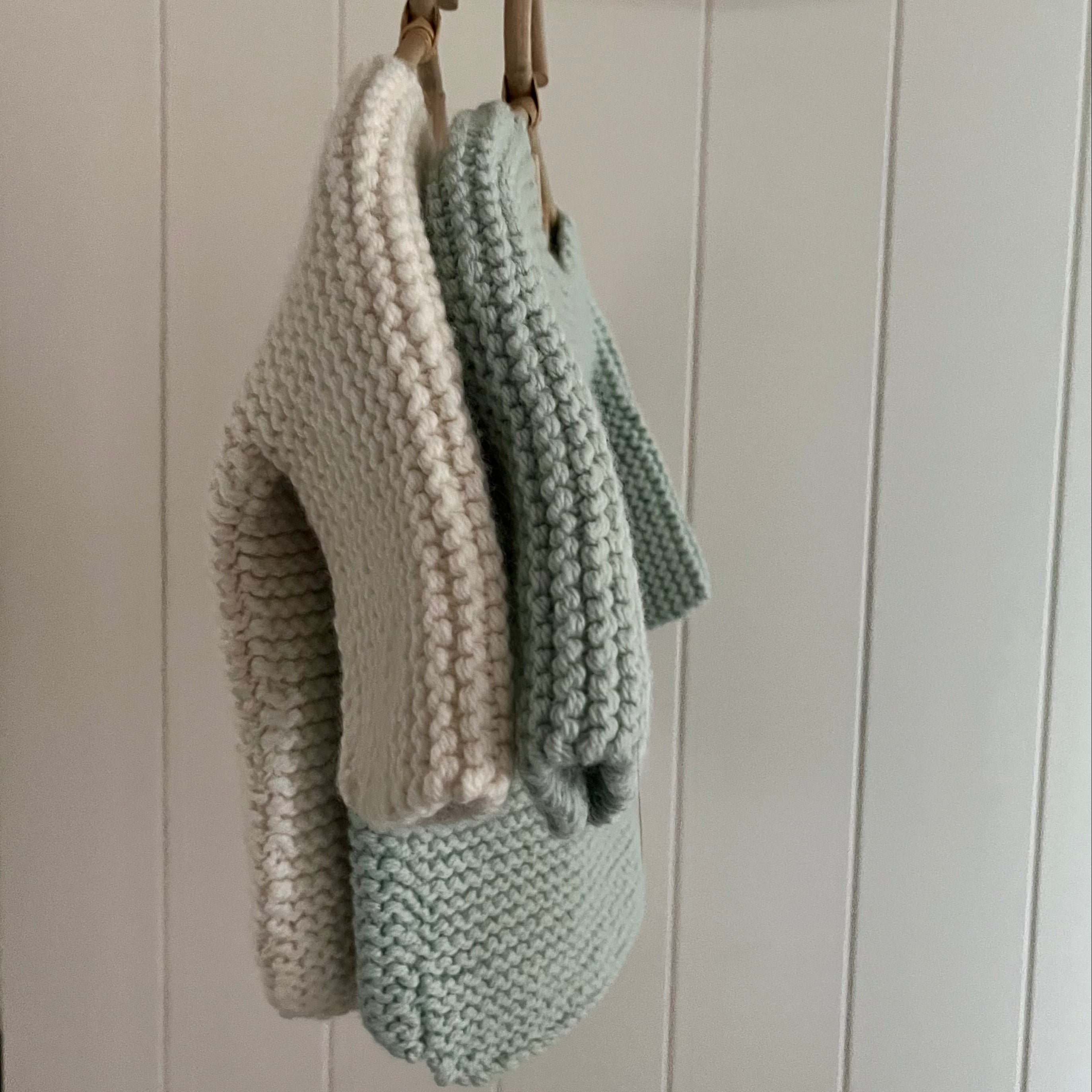 Two knitted items, one natural and one green, hanging on a hook against a white paneled wall.
