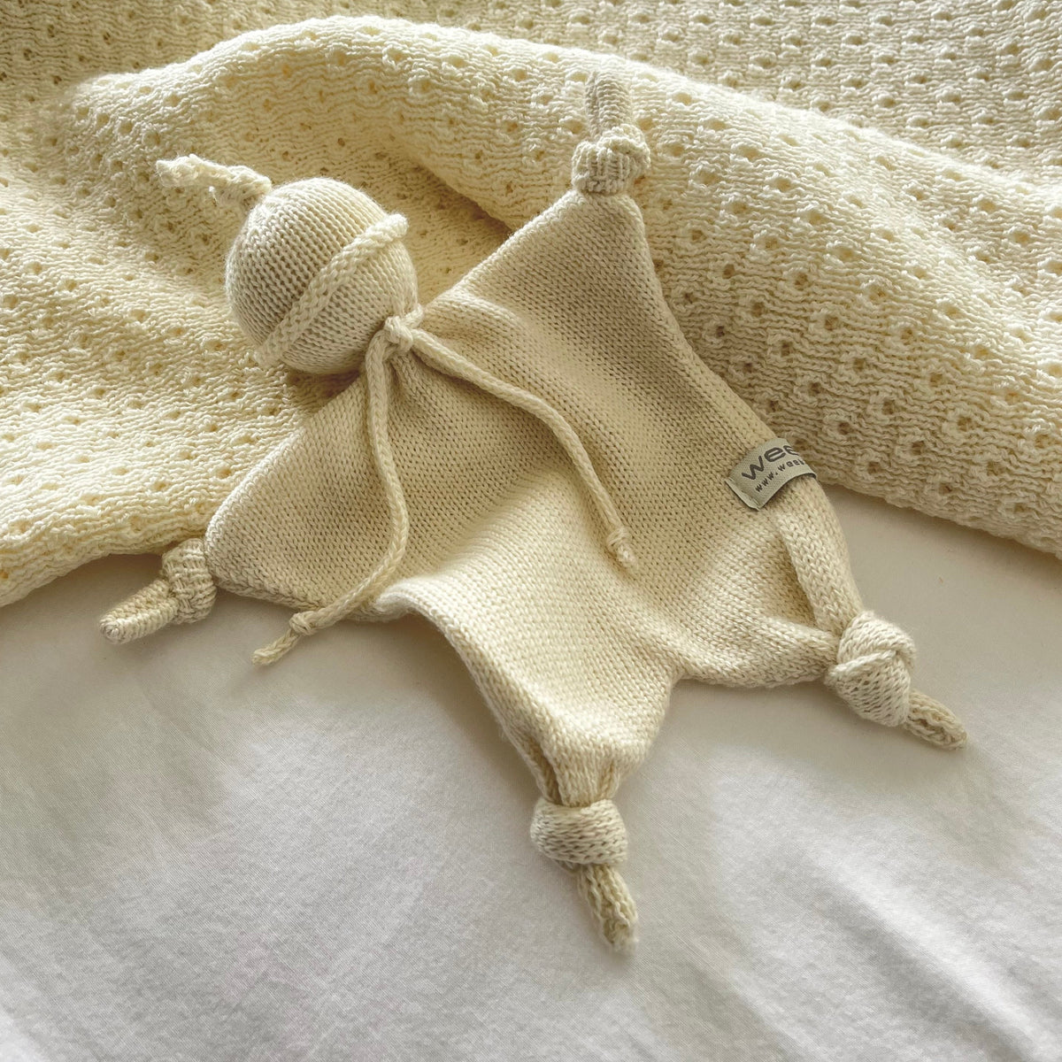 A cream-colored knitted baby lovey security blanket with a knitted ball head and four knotted tassels, styled on a white sheet with a textured cream blanket in the background.