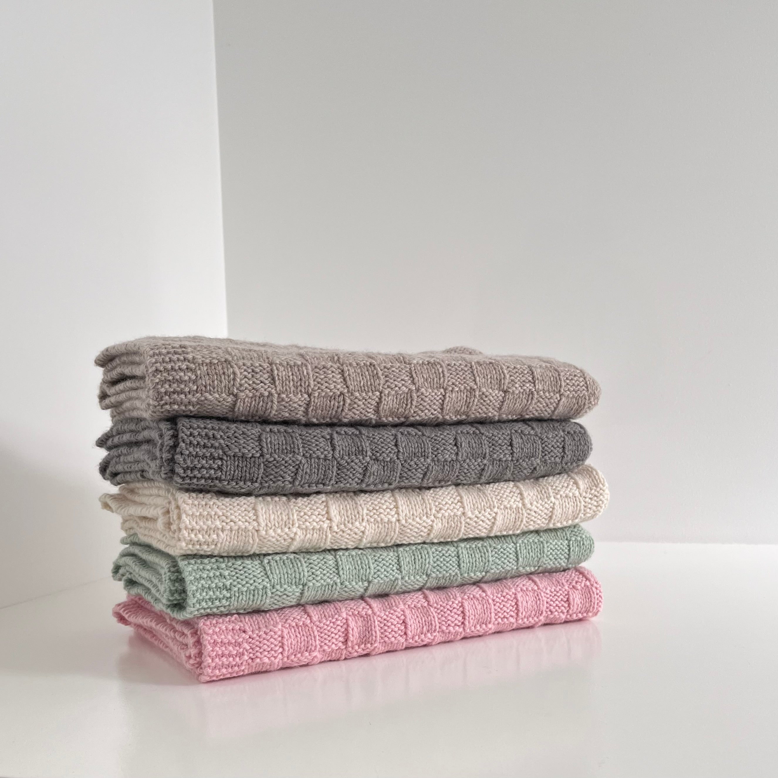 Stack of folded baby basket weave blankets in gray, natural, green, and pink on a light background