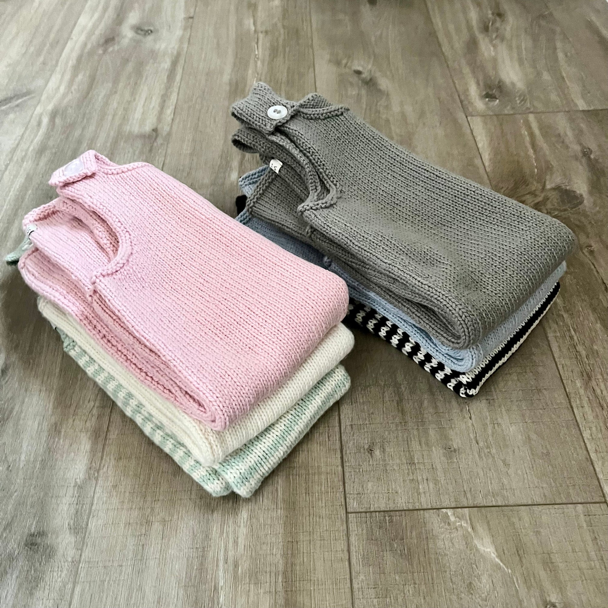Folded knit baby garments stacked on a wood floor: a light pink dungaree with a small white button closure on top, and a mushroom knit piece on top
