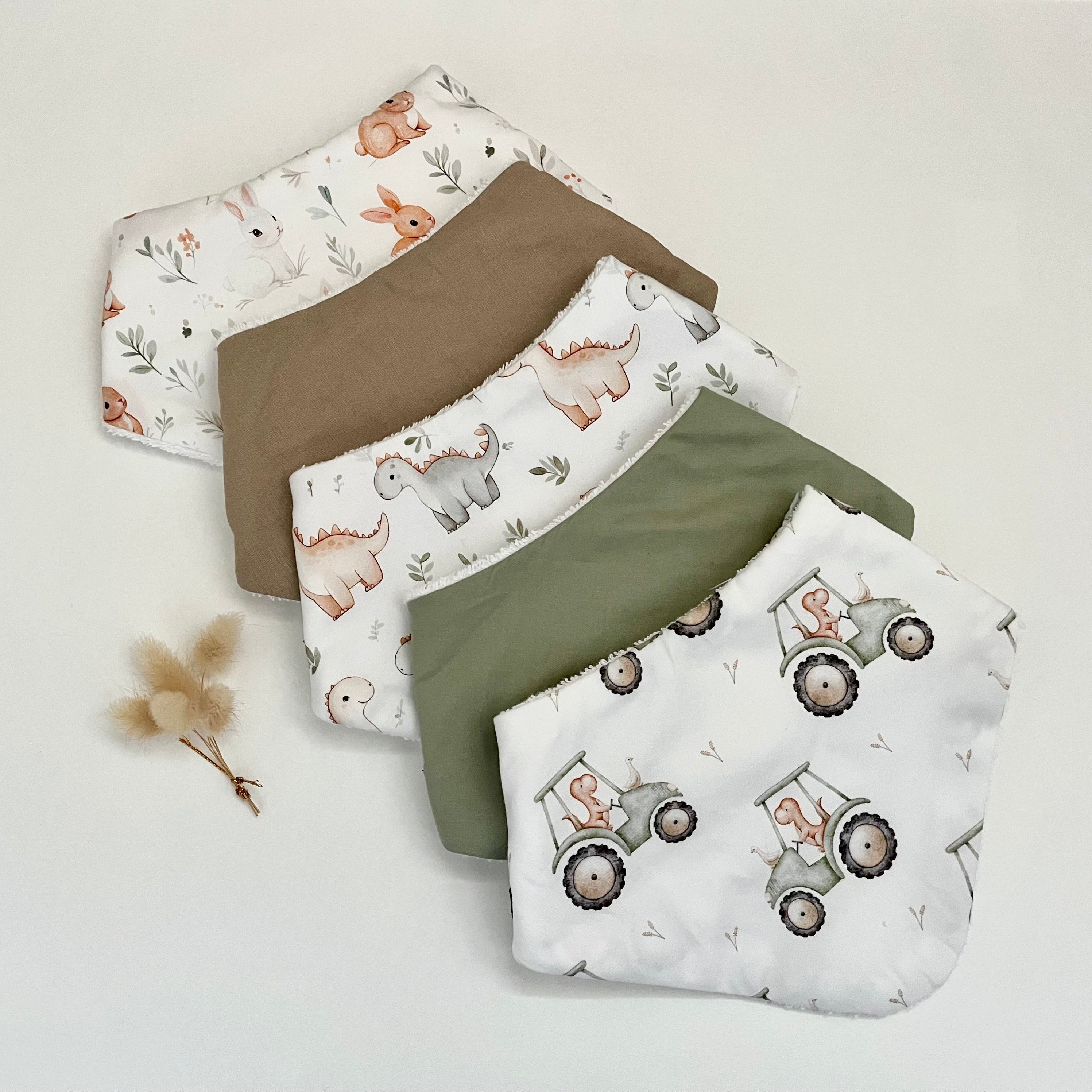 Five reusable cloth bibs arranged in a fan shape on a light neutral background. The set includes two solid brown pieces, two white bibs with animal prints (a rabbit and a dinosaur), and one solid olive green bib.