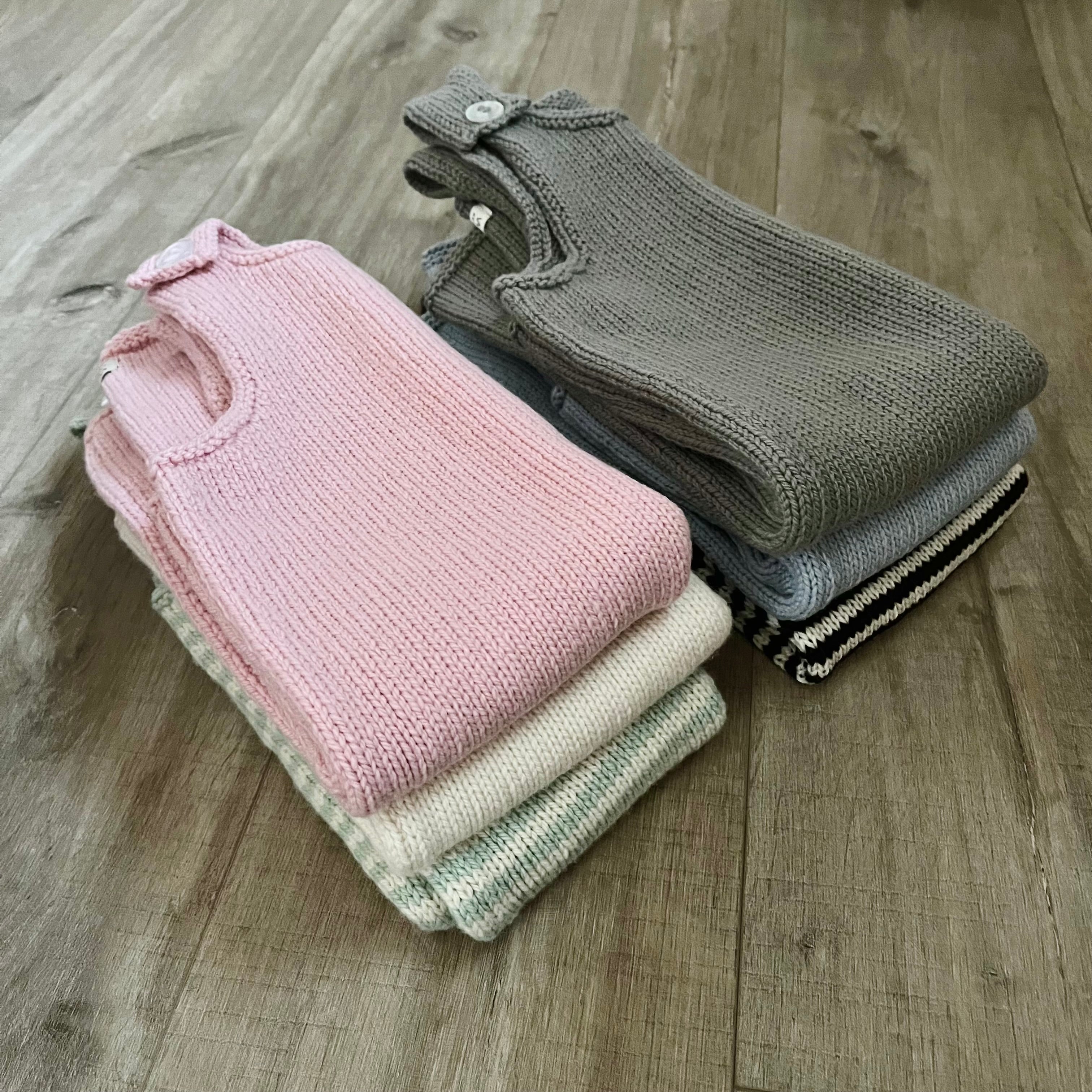 Three folded knit baby dungarees in pink, gray, and cream are stacked on a light brown wooden floor.