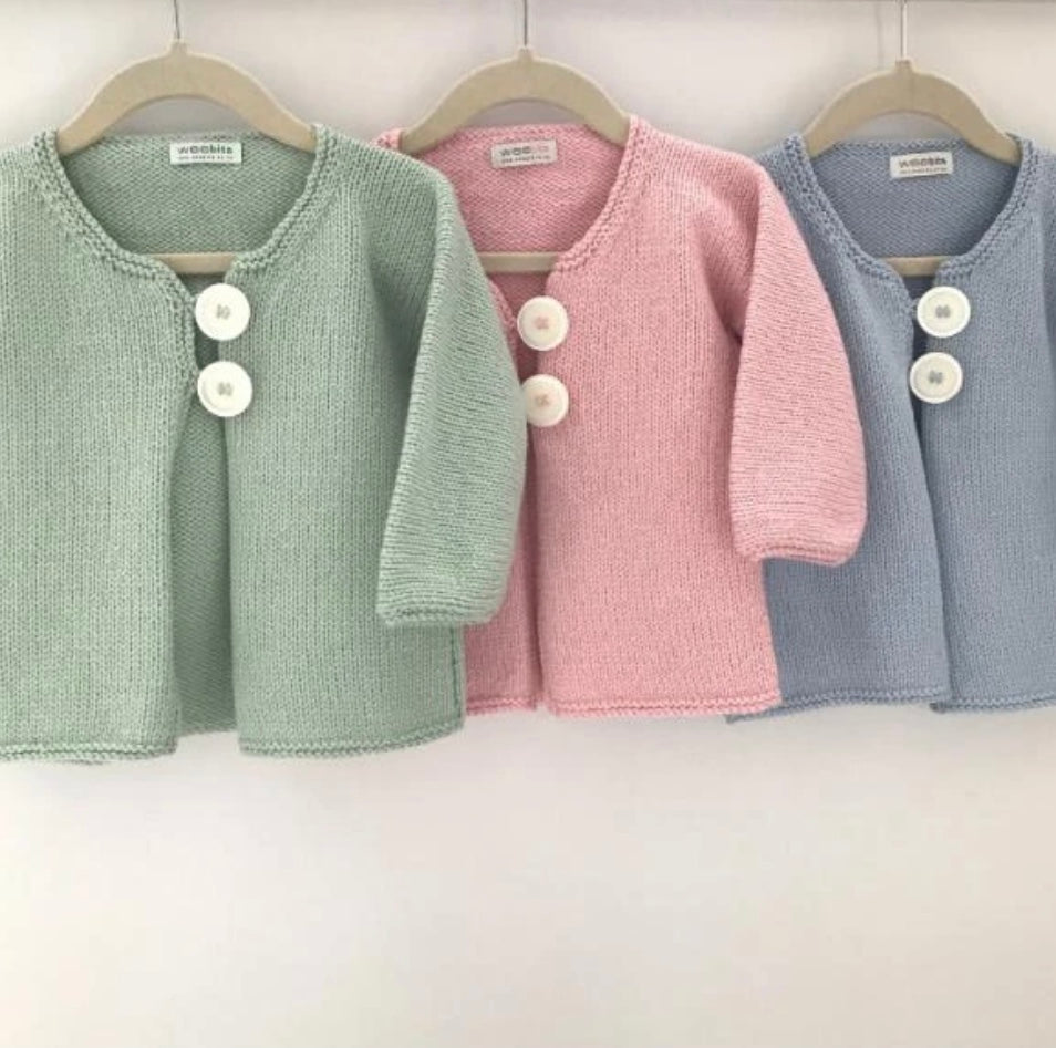 Three knitted baby cardigans in sage green, dusty pink, and light blue, hanging on wooden hangers against a plain white wall. Each cardigan features a round neckline, two white buttons, and short sleeves.