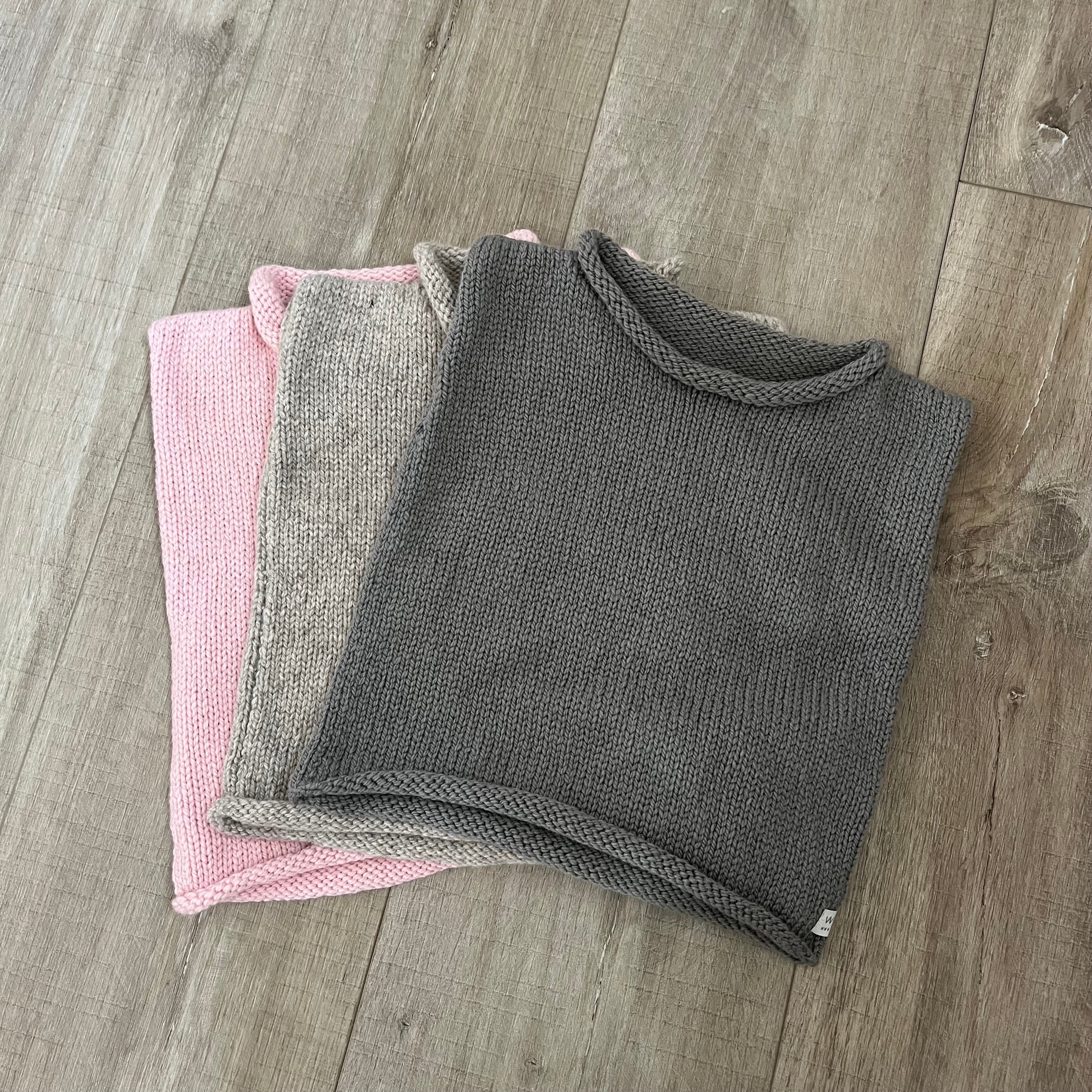 Three knit vests in gray, beige, and pink are stacked on a light brown wood floor.