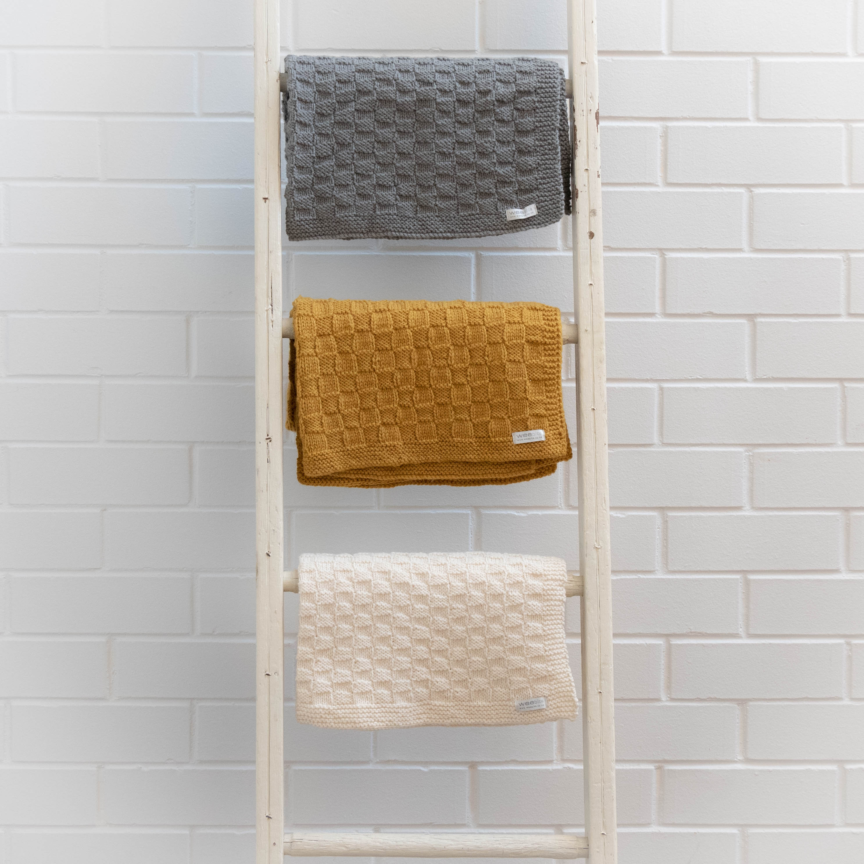 Ladder with three textured rugs in gray, mustard, and natural against a white tiled wall.