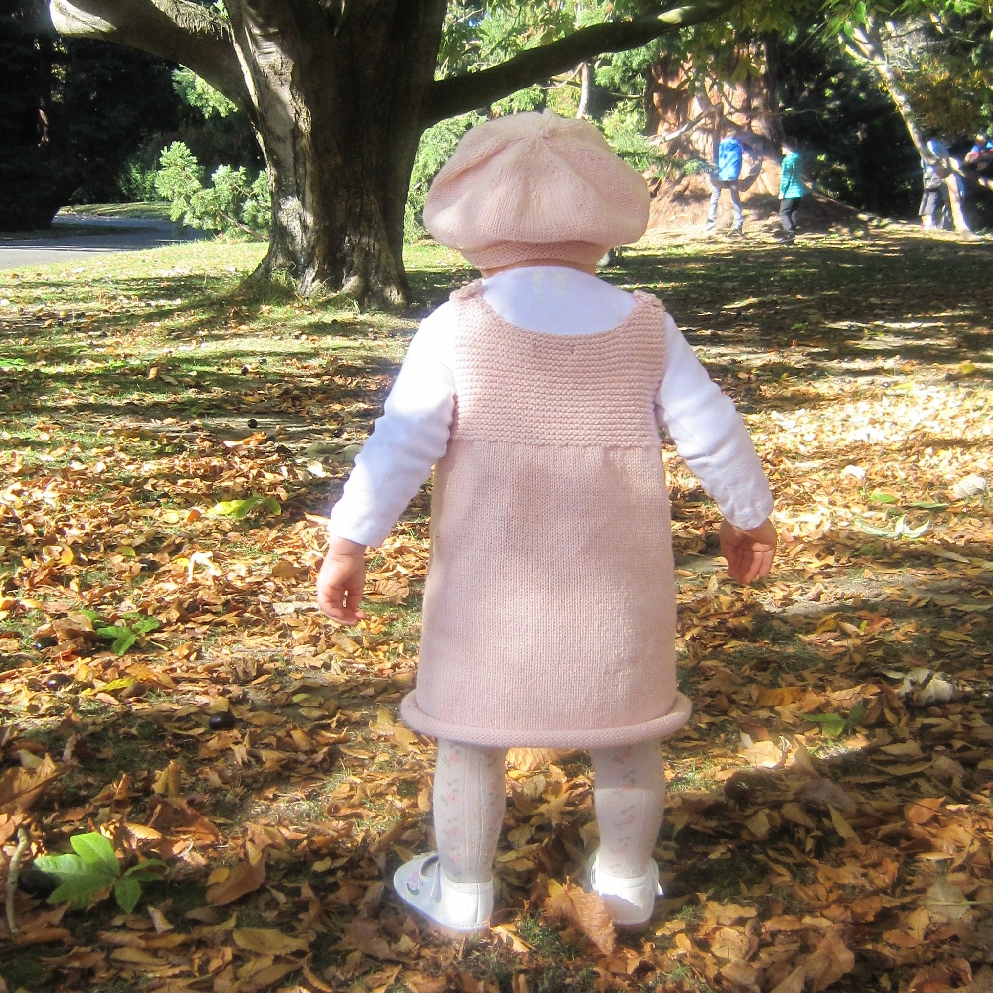 A baby wearing a light pink knit dress, white long-sleeve shirt, matching beret, and white booties, standing on a leaf-covered ground in a park.