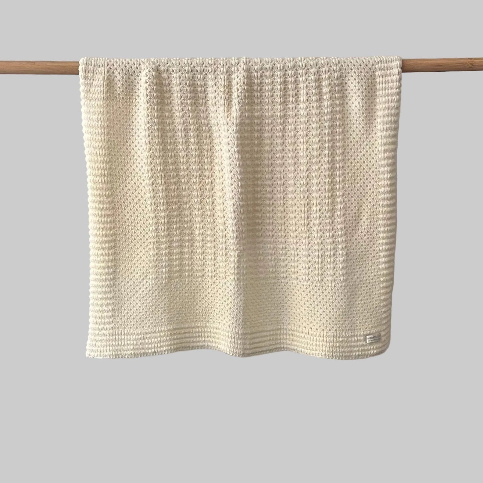 Textured buttermilk bassinet blanket hanging on a wooden rod against a grey background
