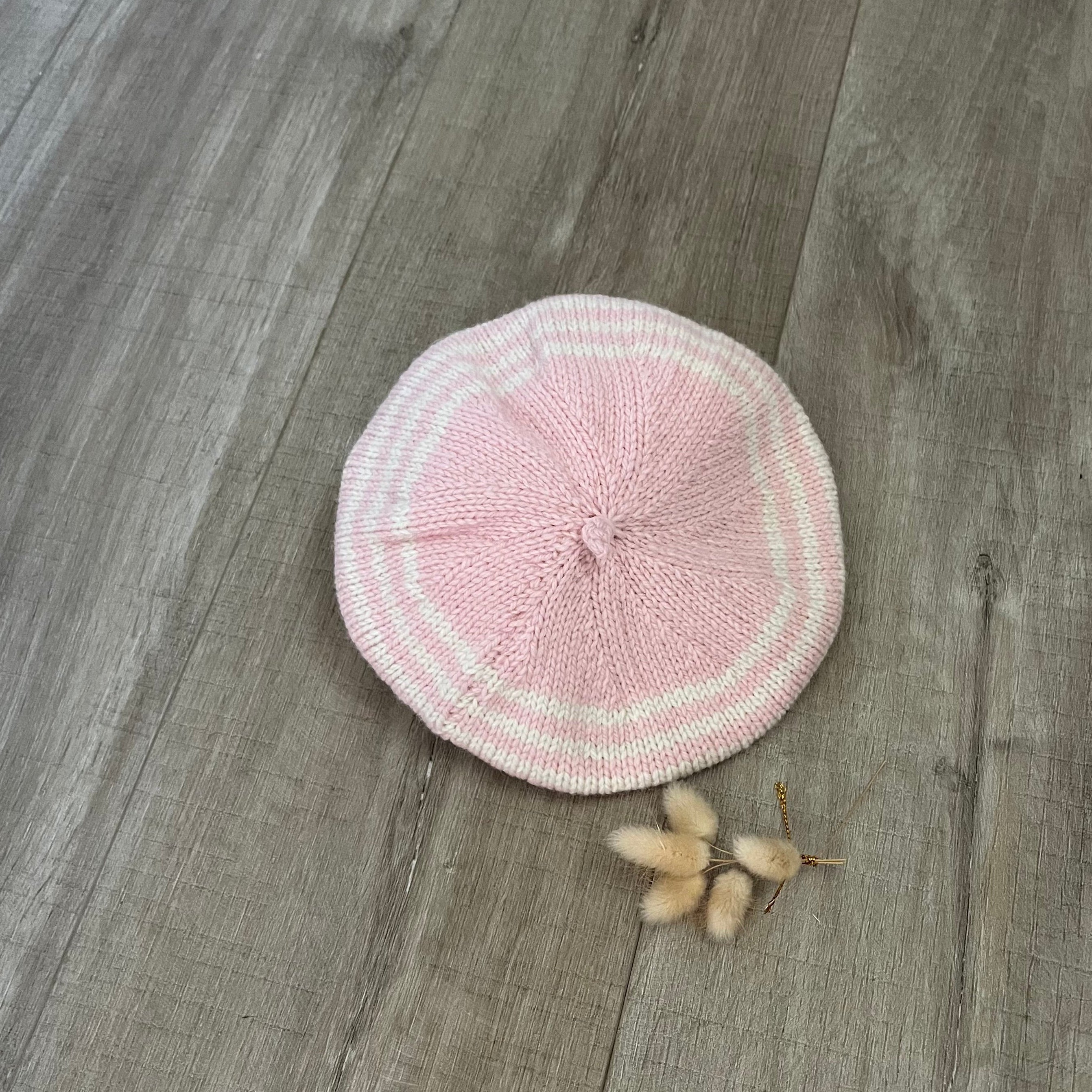 Pink stripe knitted beret on a wooden surface with dried flowers.