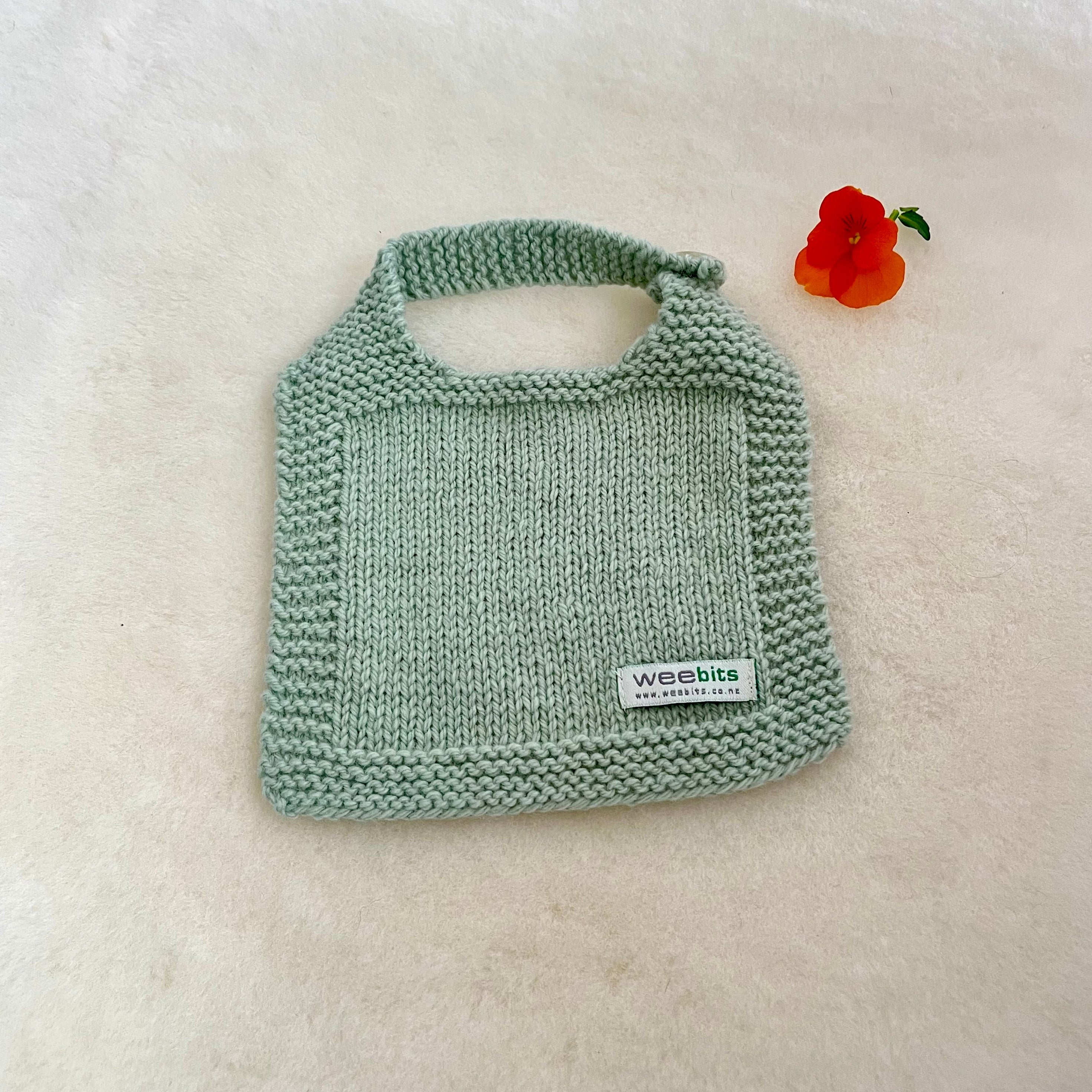 A light mint green hand-knit baby bib with a small white brand label reading 'weebis', displayed on a soft white fur surface next to a small orange flower.