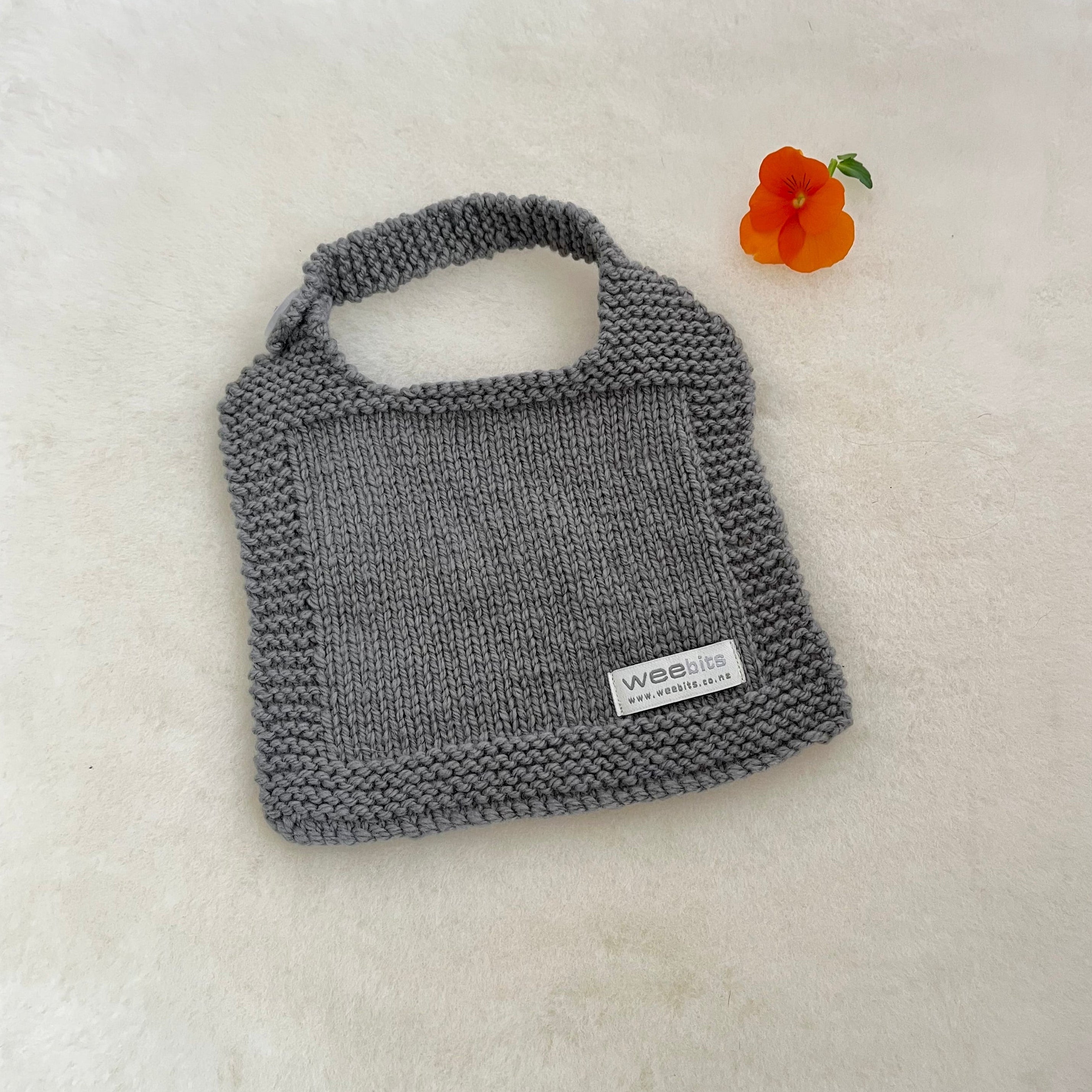 A gray hand-knitted baby bib laid flat on a white textured surface next to a small orange flower. A white brand label reading 'weebits' sewn onto the bottom right corner.