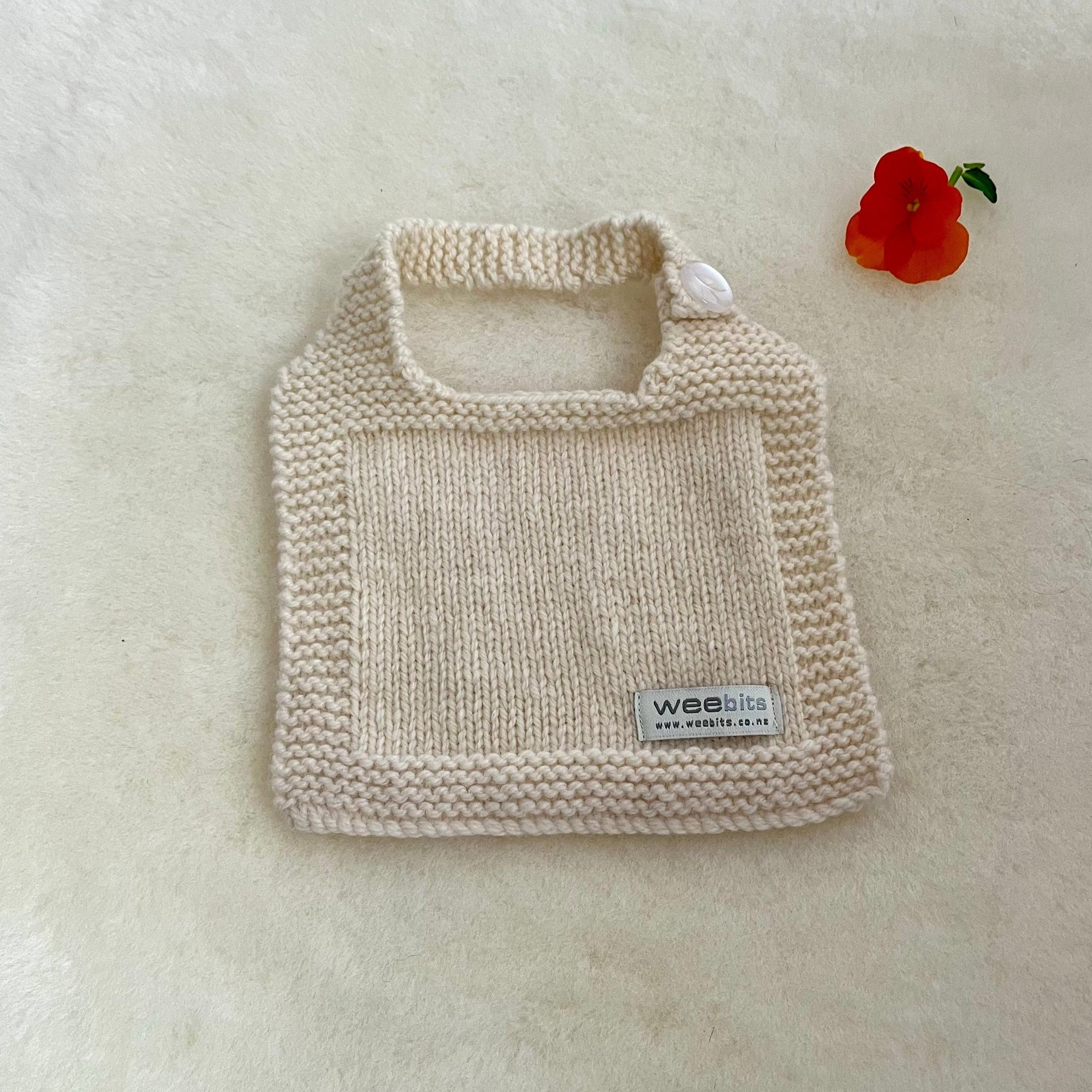 A cream-colored hand-knit baby bib displayed on a soft white textured background with a small red flower prop in the corner.