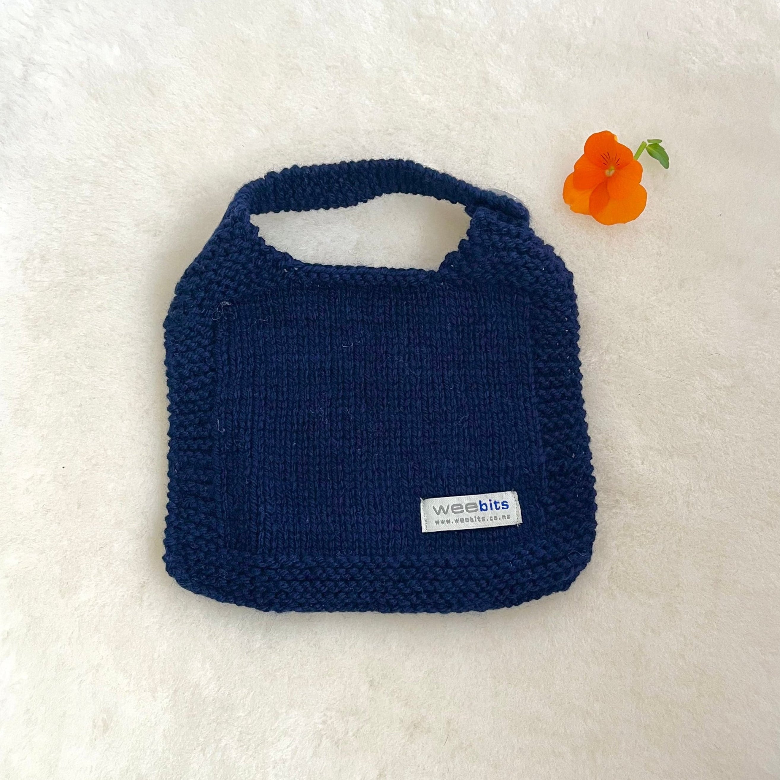 A navy blue hand-knit baby bib with a small white 'weebits' brand label, laid flat on a white textured background next to a small orange flower.